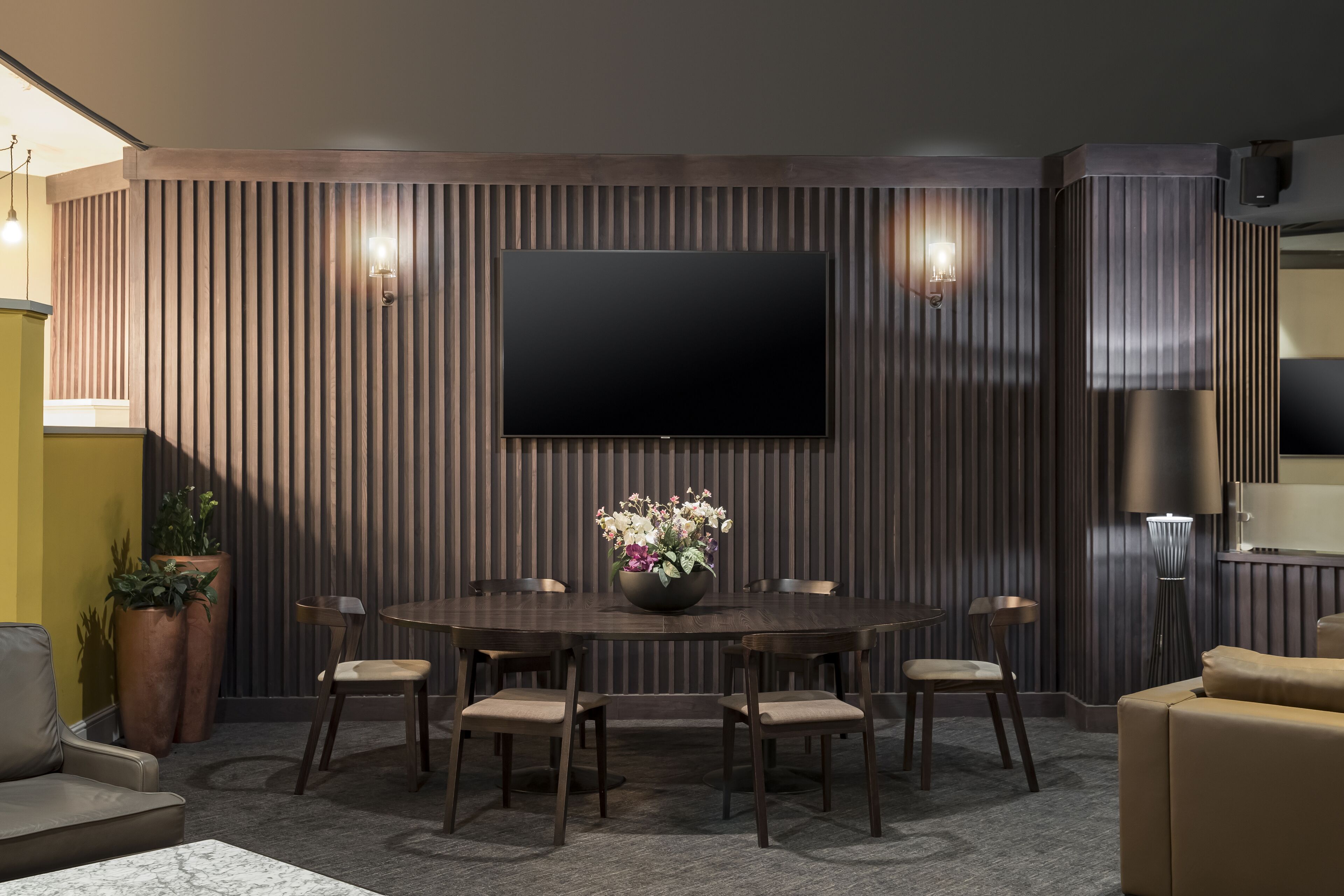 A modern dining area with a round table, six chairs, a flat-screen TV mounted on a wooden wall, and a potted plant with flowers.