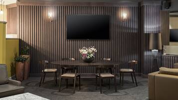 A modern dining area with a round table, six chairs, a flat-screen TV mounted on a wooden wall, and a potted plant with flowers.