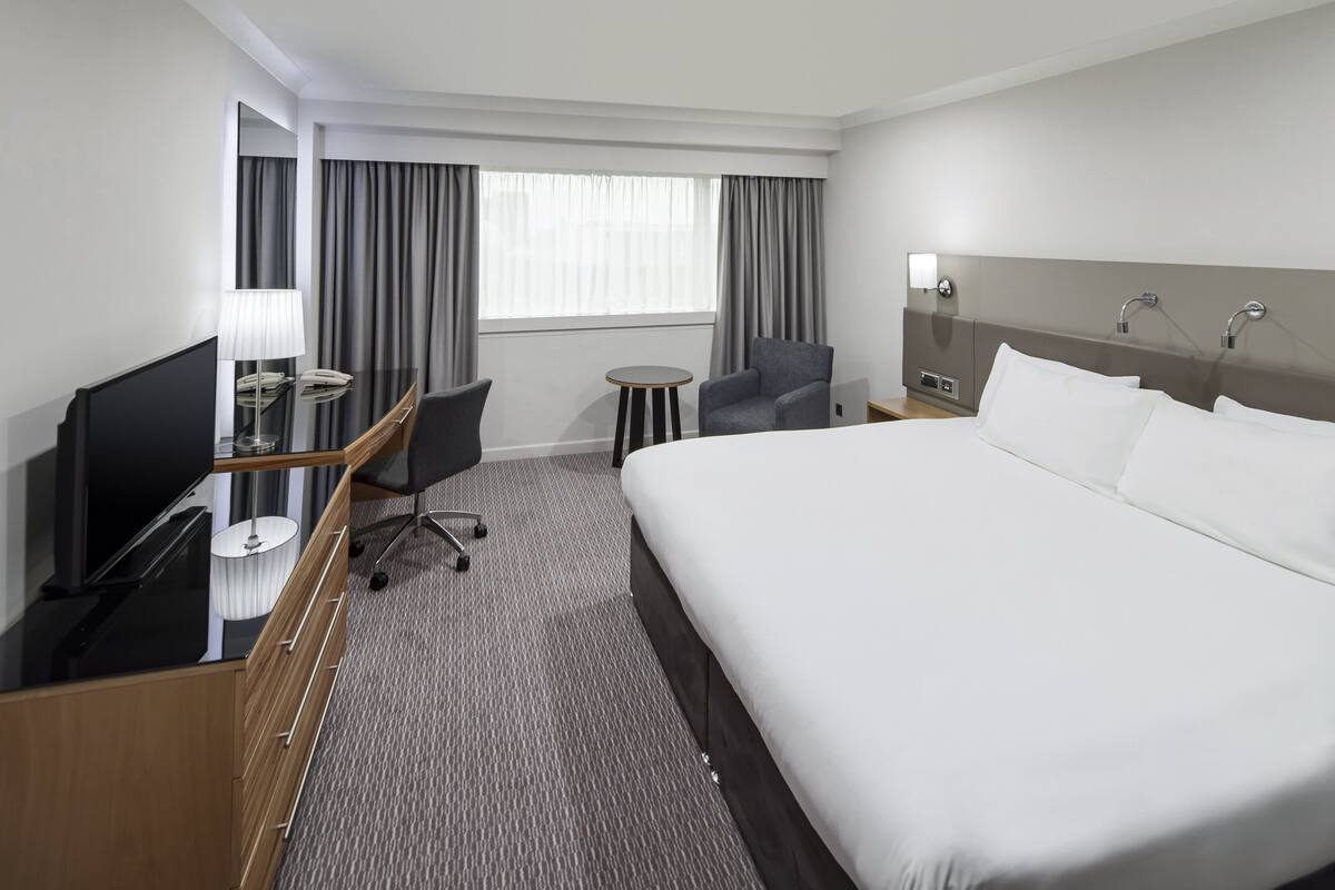 A hotel room with a large bed, a desk with a TV, a chair, a small table, and a window with curtains.