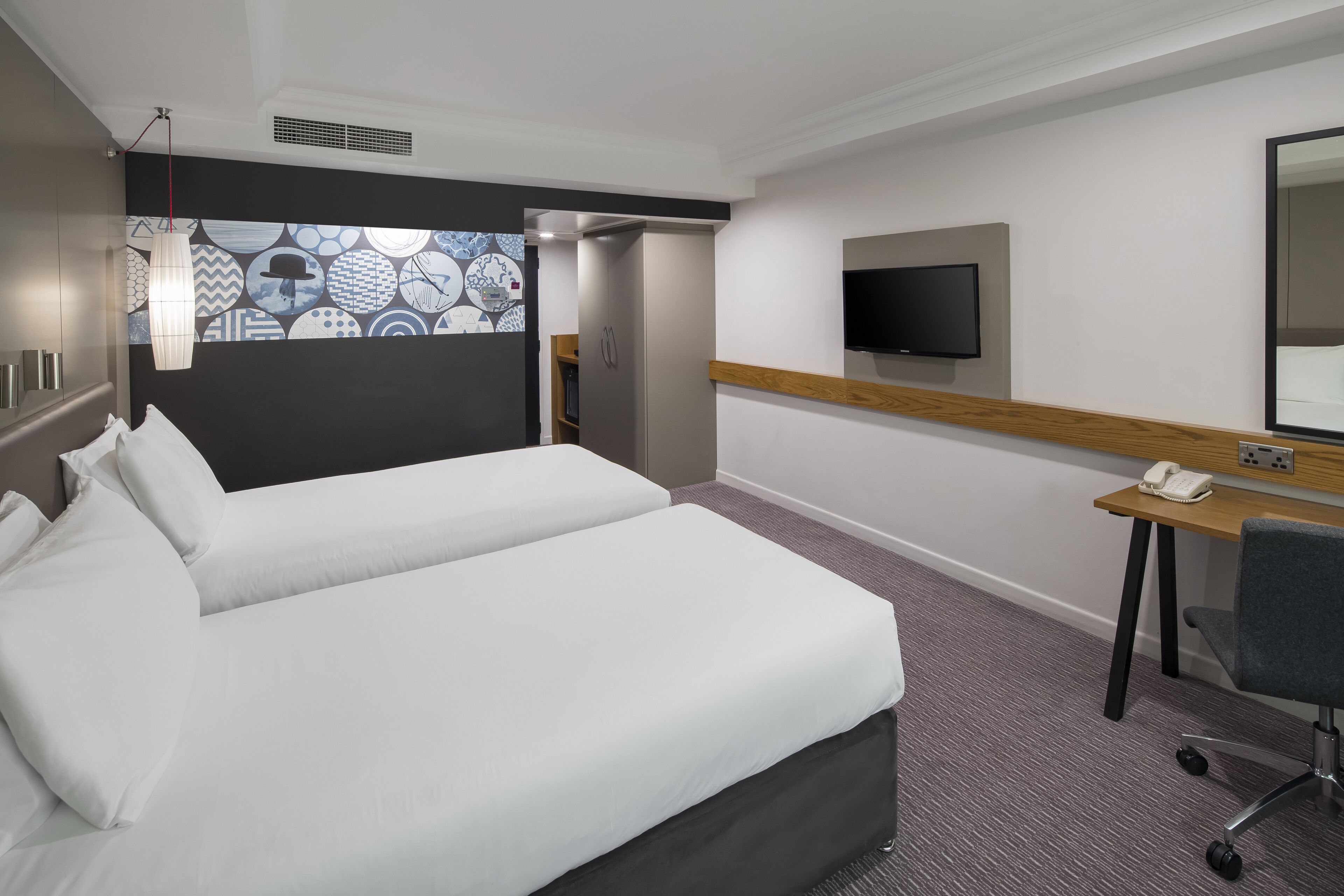 A hotel room with a large bed, a desk with a phone, a TV, and a patterned wall.