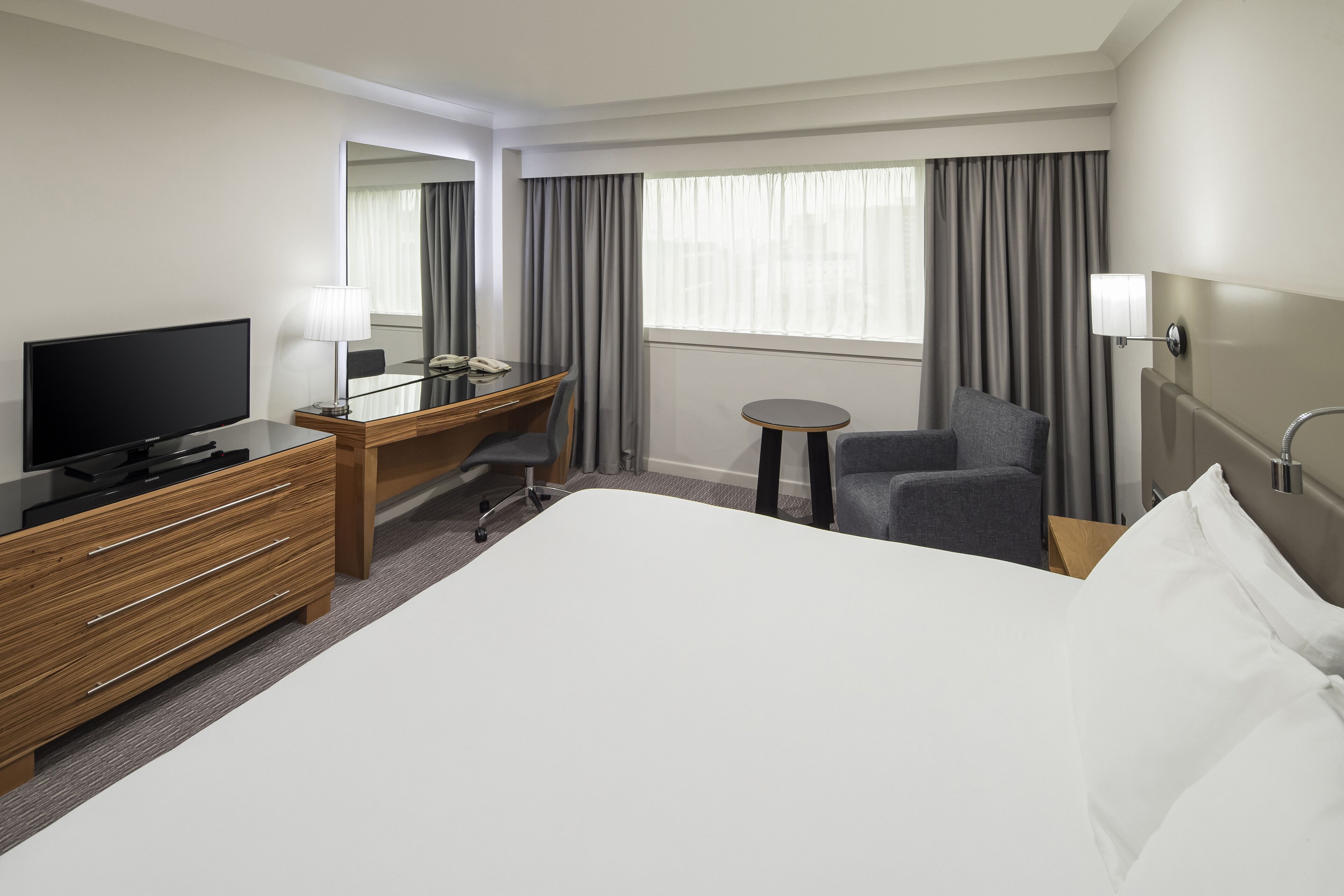 A hotel room with a large bed, a desk with a television, a chair, a small table, and a window with curtains.