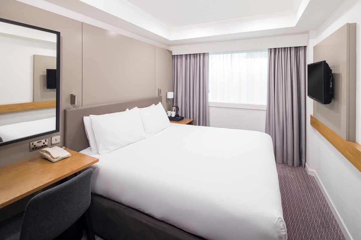 Ein Hotelzimmer mit einem großen Bett, einem Schreibtisch mit Telefon, einem Fernseher und einem Fenster mit Vorhängen.