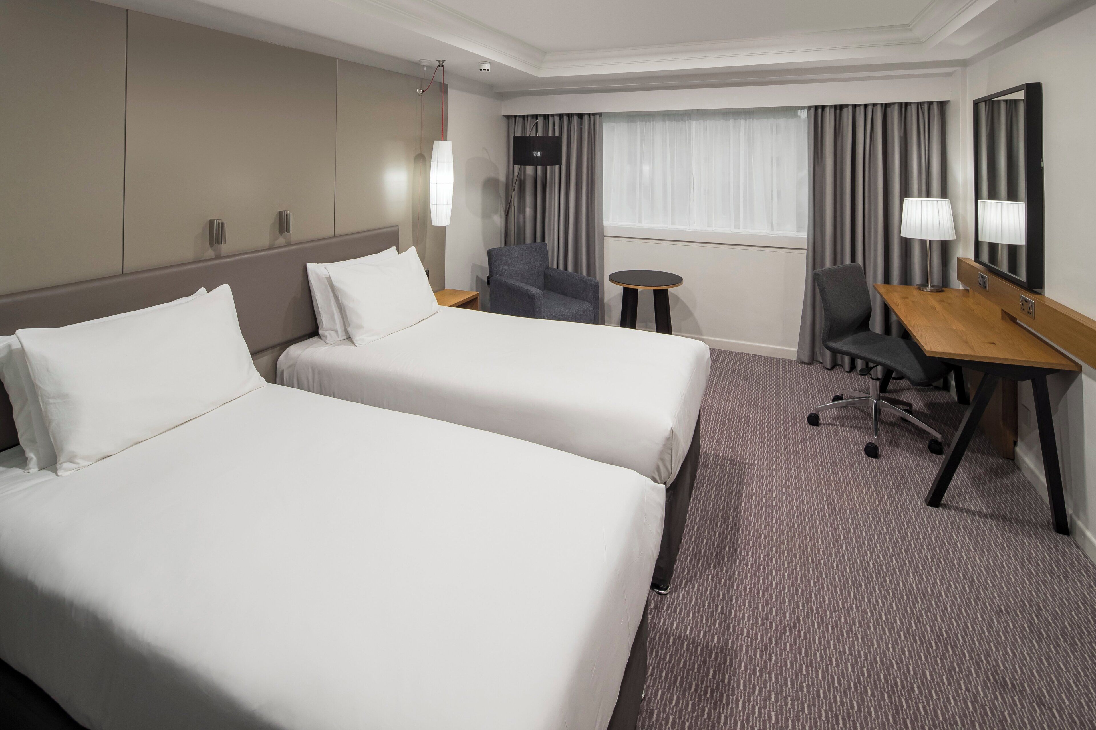 Habitación de hotel con dos camas, un escritorio, una silla, una mesita y una ventana con cortinas.