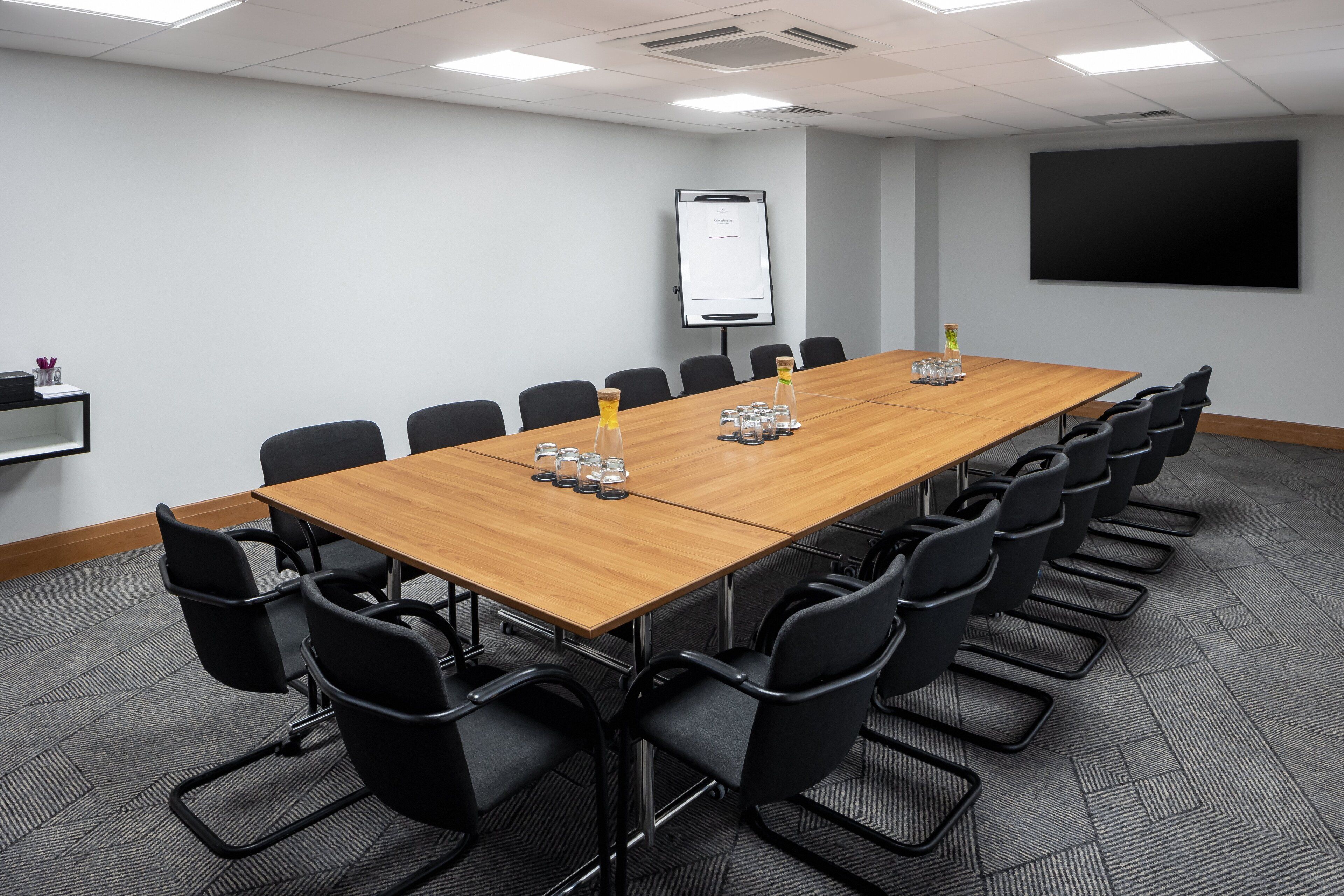 A conference room with a long wooden table, black chairs, a whiteboard, and a projector screen.