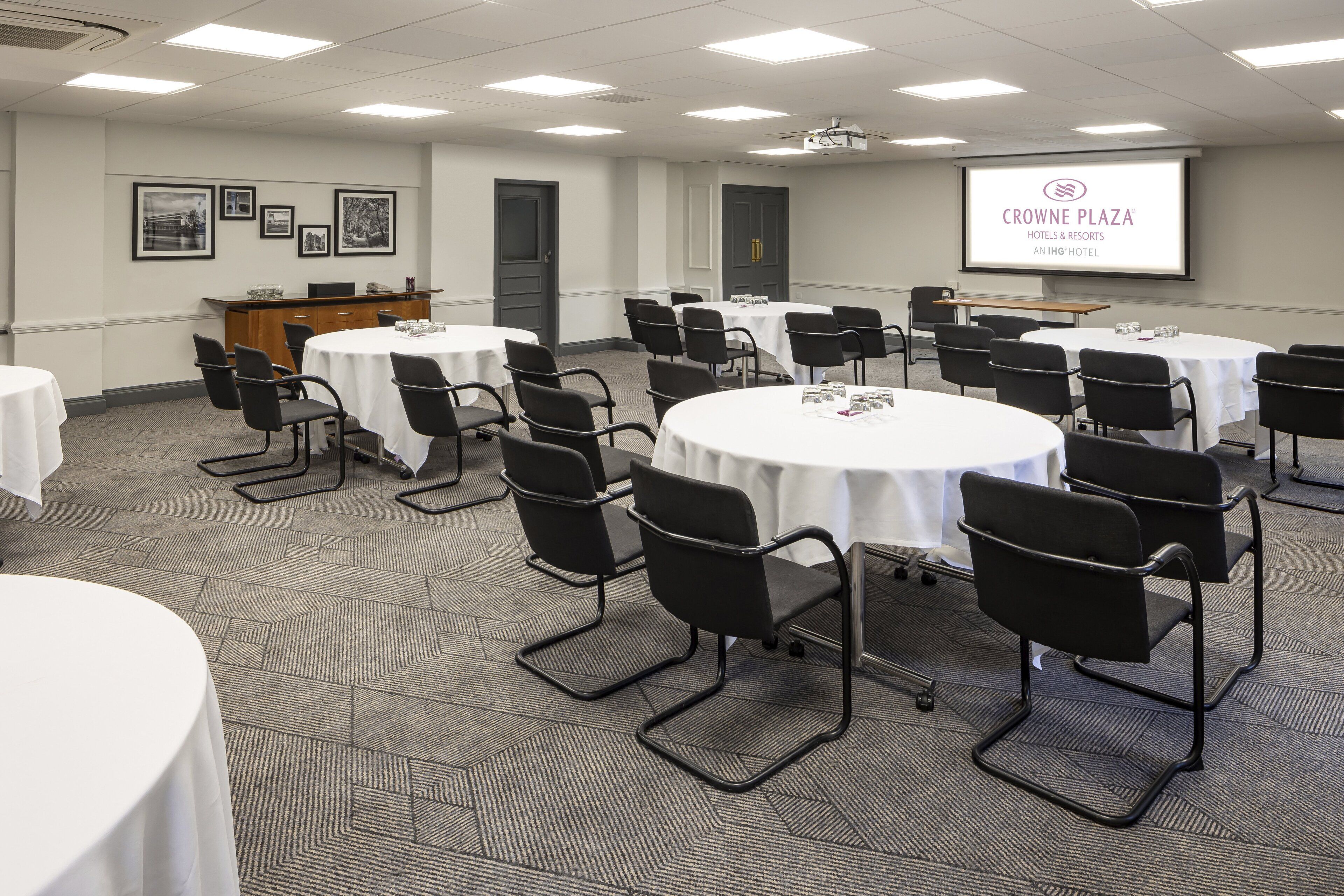 A conference room with multiple tables and chairs, a projector screen displaying 'CROWNE PLAZA HOTELS & RESORTS AN IHG HOTEL', and framed pictures on the wall.