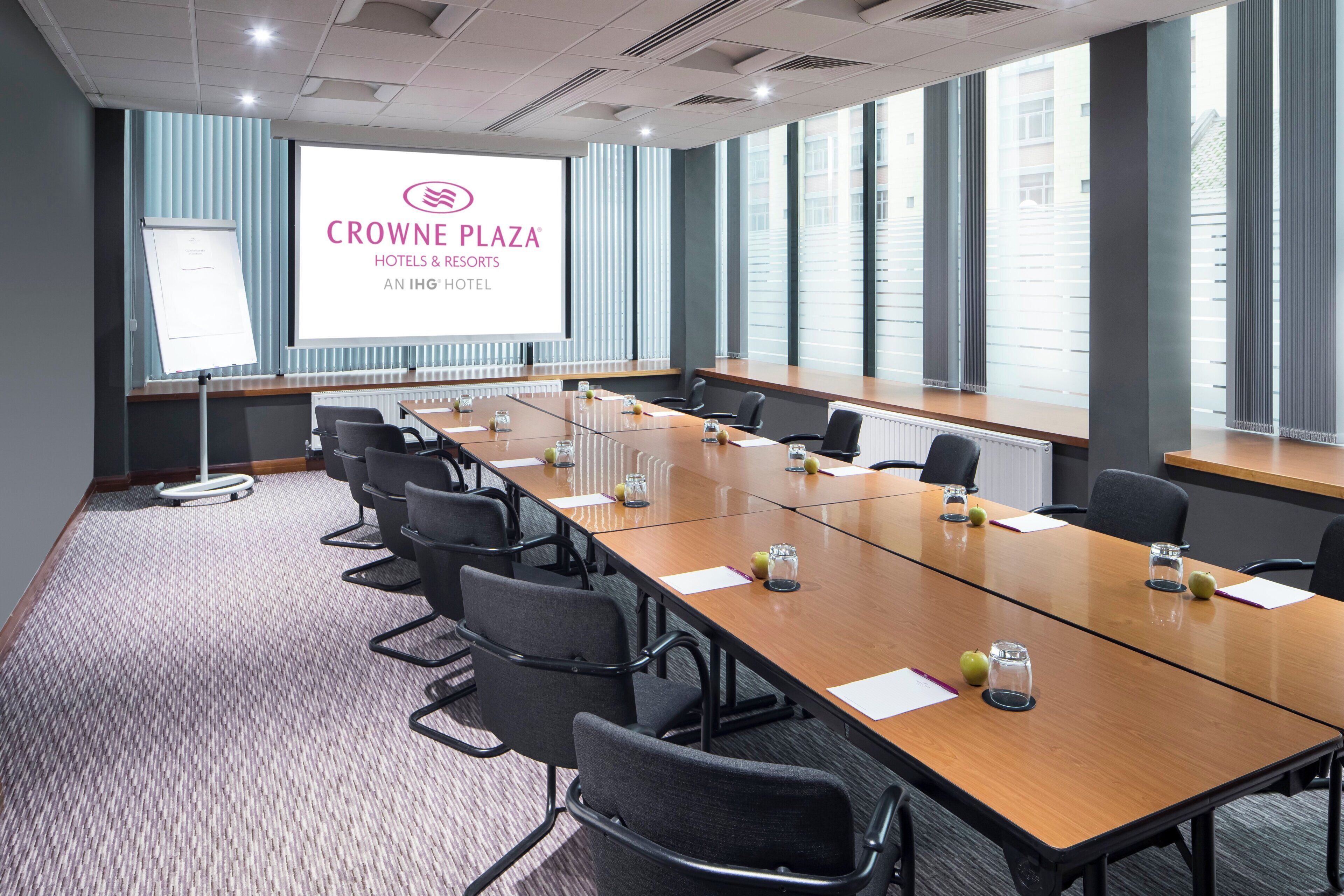 A conference room with a long table, chairs, a projector screen displaying 'CROWNE PLAZA HOTELS & RESORTS AN IHG HOTEL', and large windows with blinds.