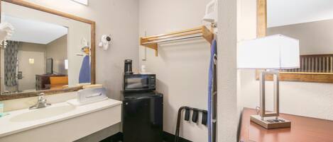 A compact hotel bathroom with a sink, mirror, microwave, and a small table with a lamp.