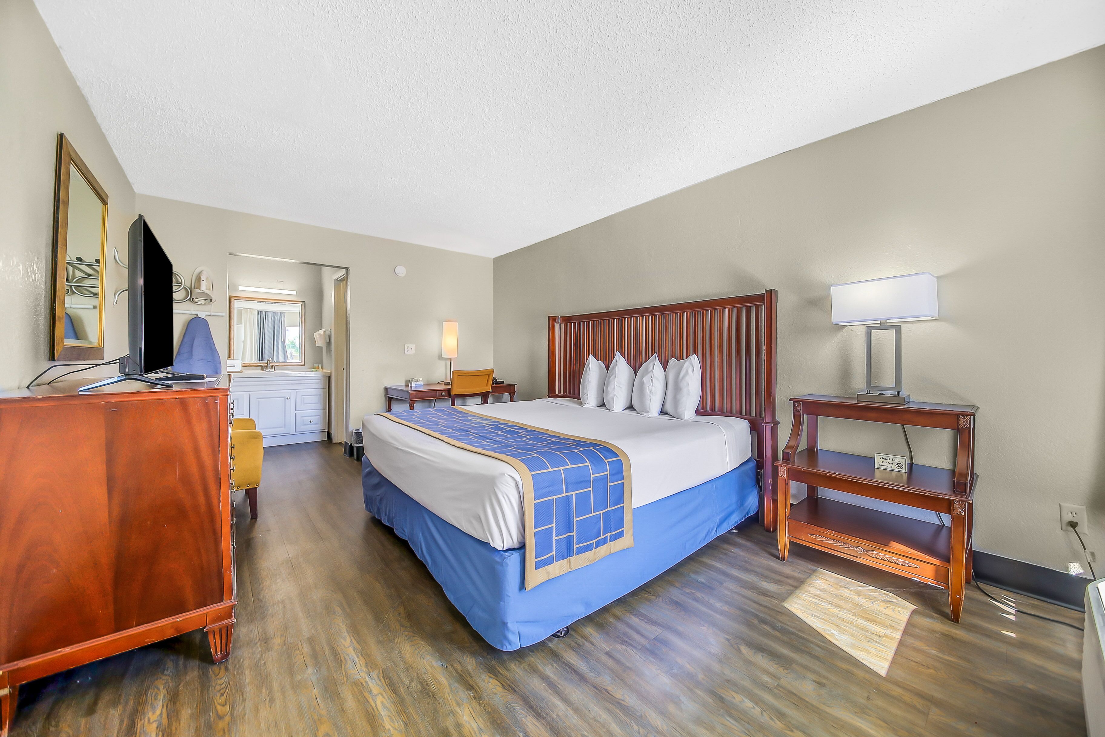 A hotel room with a large bed, a wooden dresser, a desk, and a nightstand with a lamp.