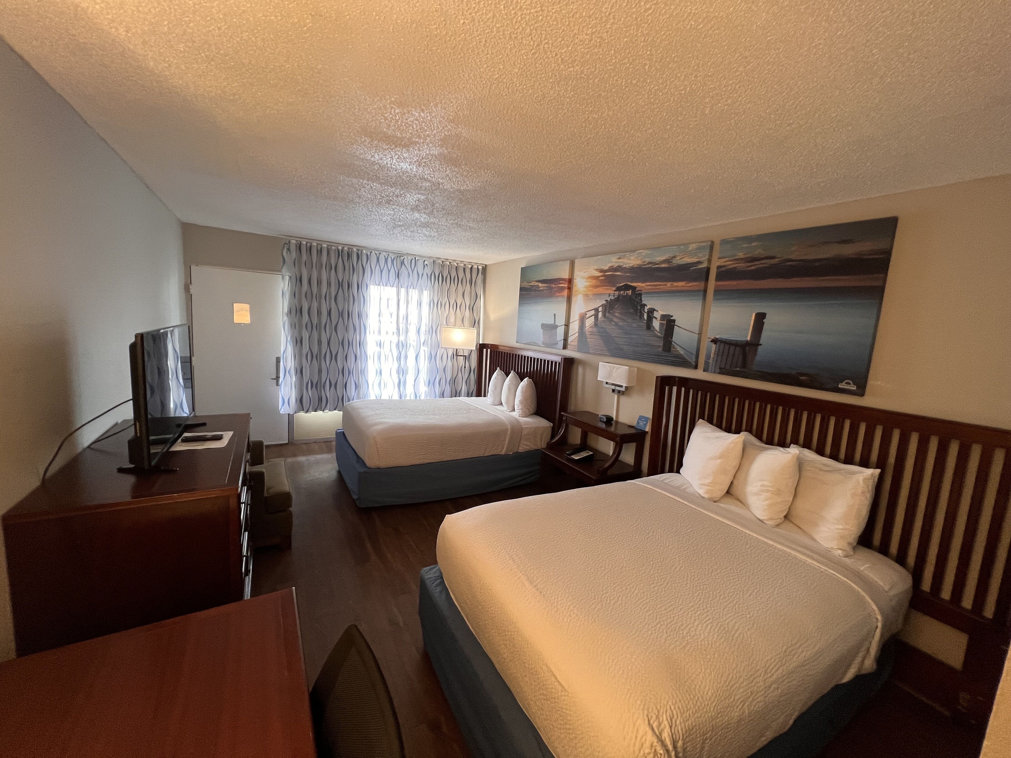 A hotel room with two beds, a desk with a TV, a lamp, and a painting of a pier.