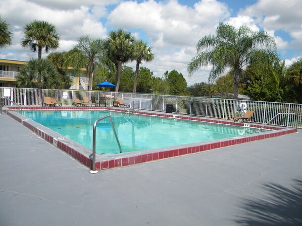 A rectangular swimming pool with a red border, surrounded by a white fence. There are lounge chairs and a blue umbrella near the pool.