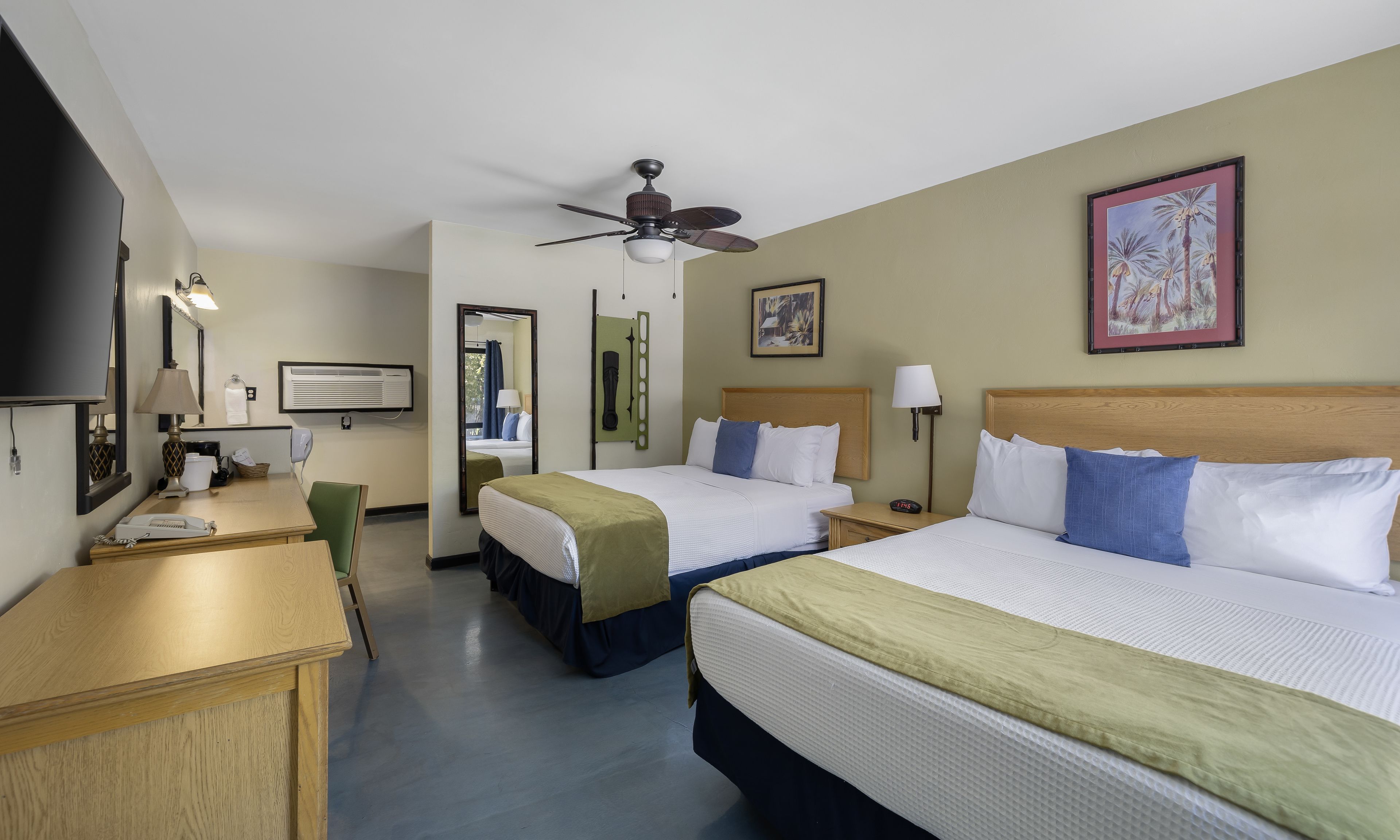 A hotel room with two beds, a desk, a chair, a ceiling fan, and a painting on the wall.