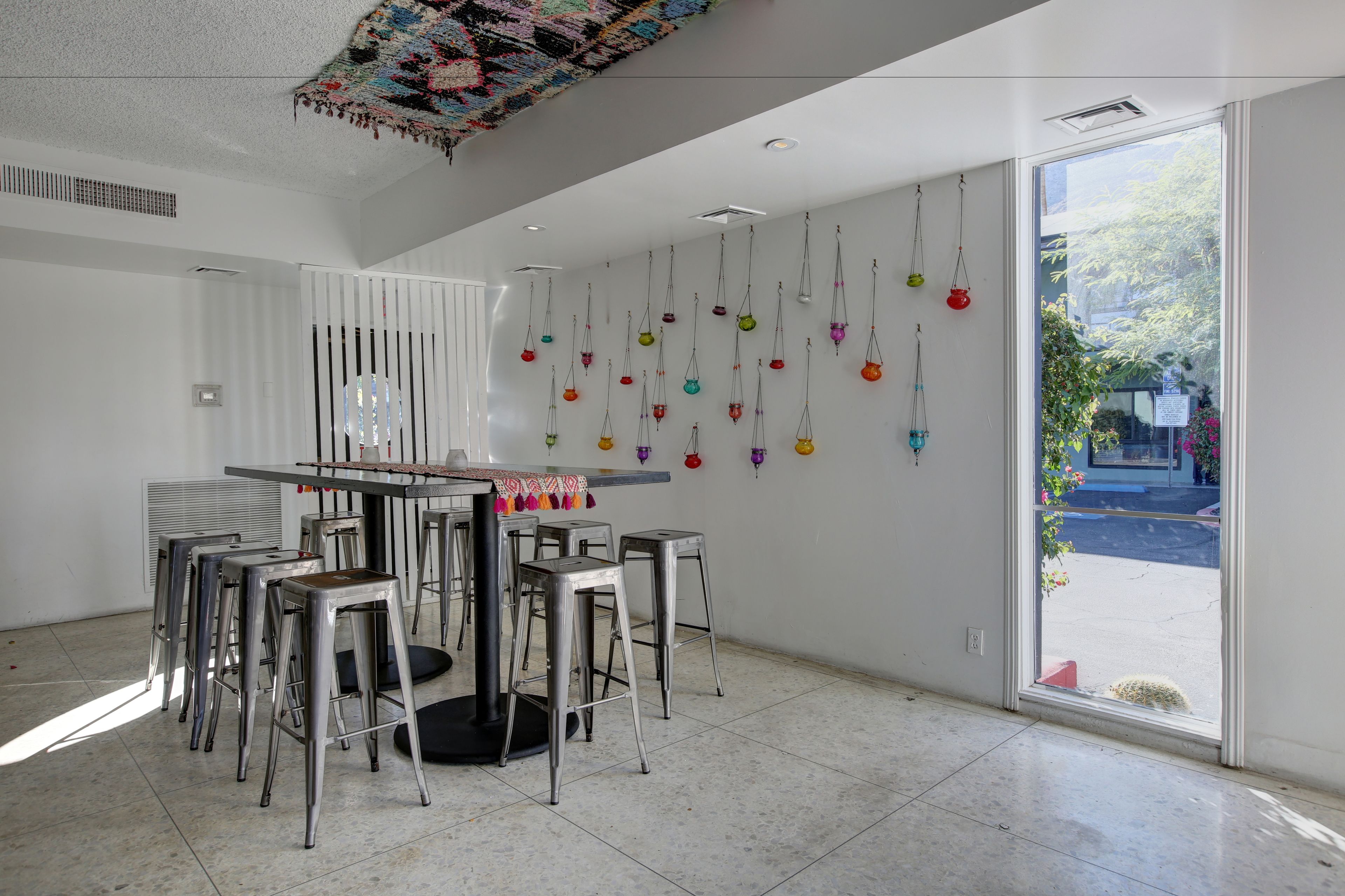A modern interior with a dining area featuring a round table and metal stools, a wall with hanging decorative items, and a large window providing a view of the outdoors.