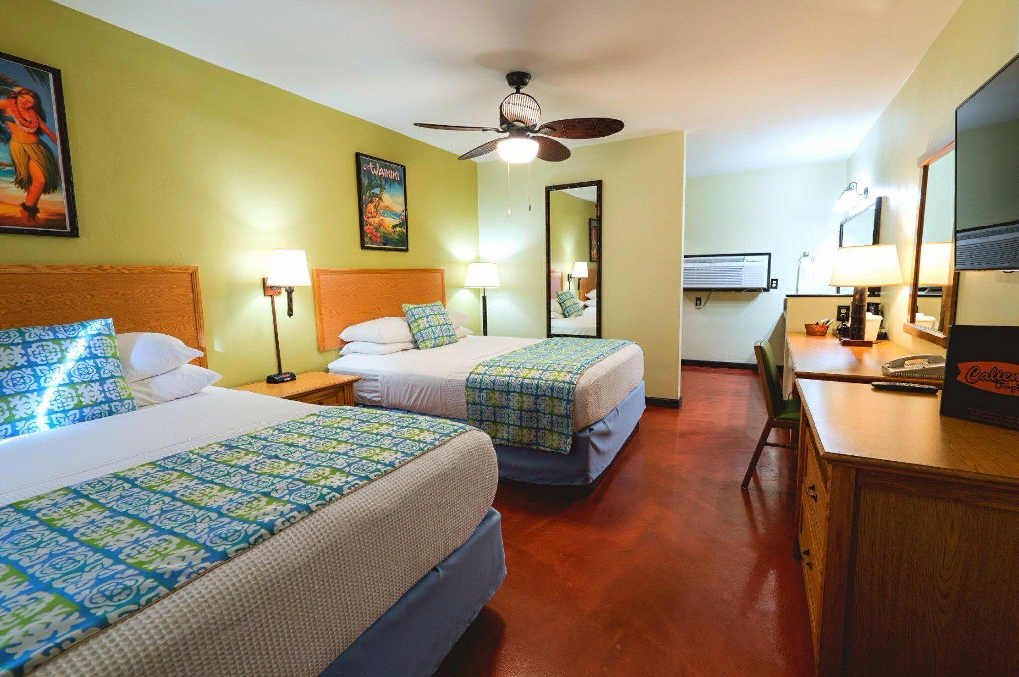 A hotel room with two beds, a desk, a chair, a ceiling fan, and a television.