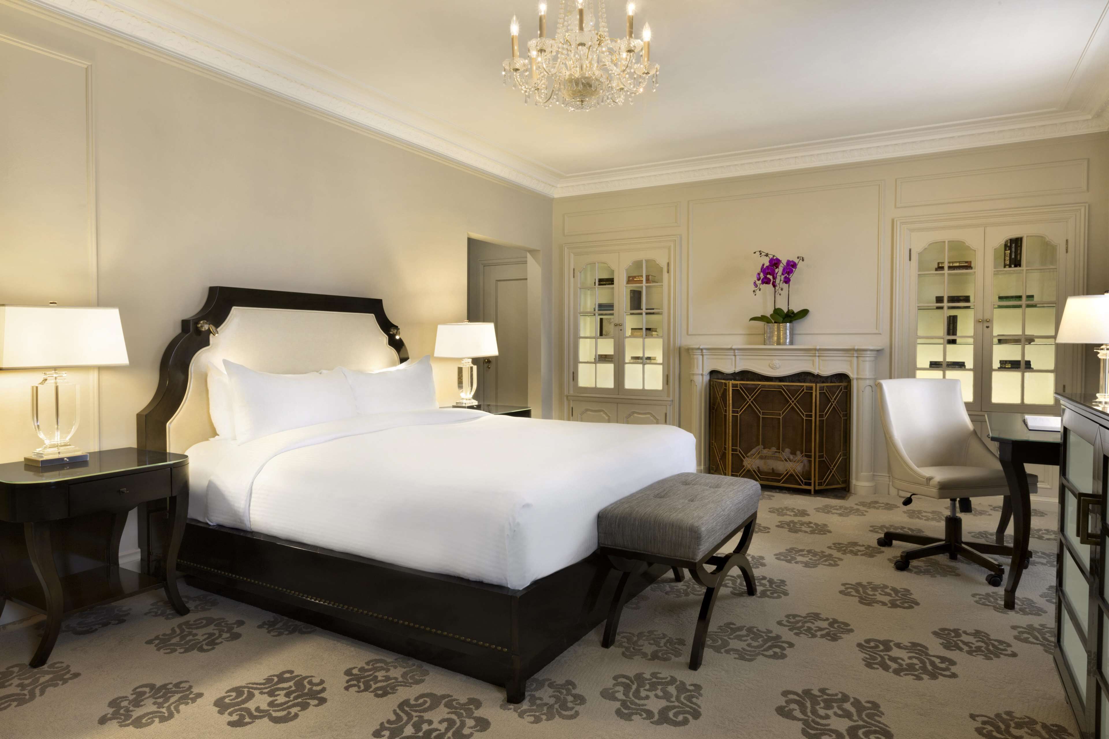 A hotel room with a large bed, two bedside tables with lamps, a chair, a desk, and a chandelier.