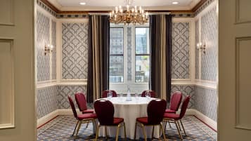 A room with a round table, red chairs, a chandelier, patterned wallpaper, and a view of buildings through large windows.