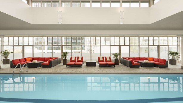 Un área de piscina interior moderna con una amplia pared de ventanas, sillones reclinables rojos y vistas despejadas del cielo.