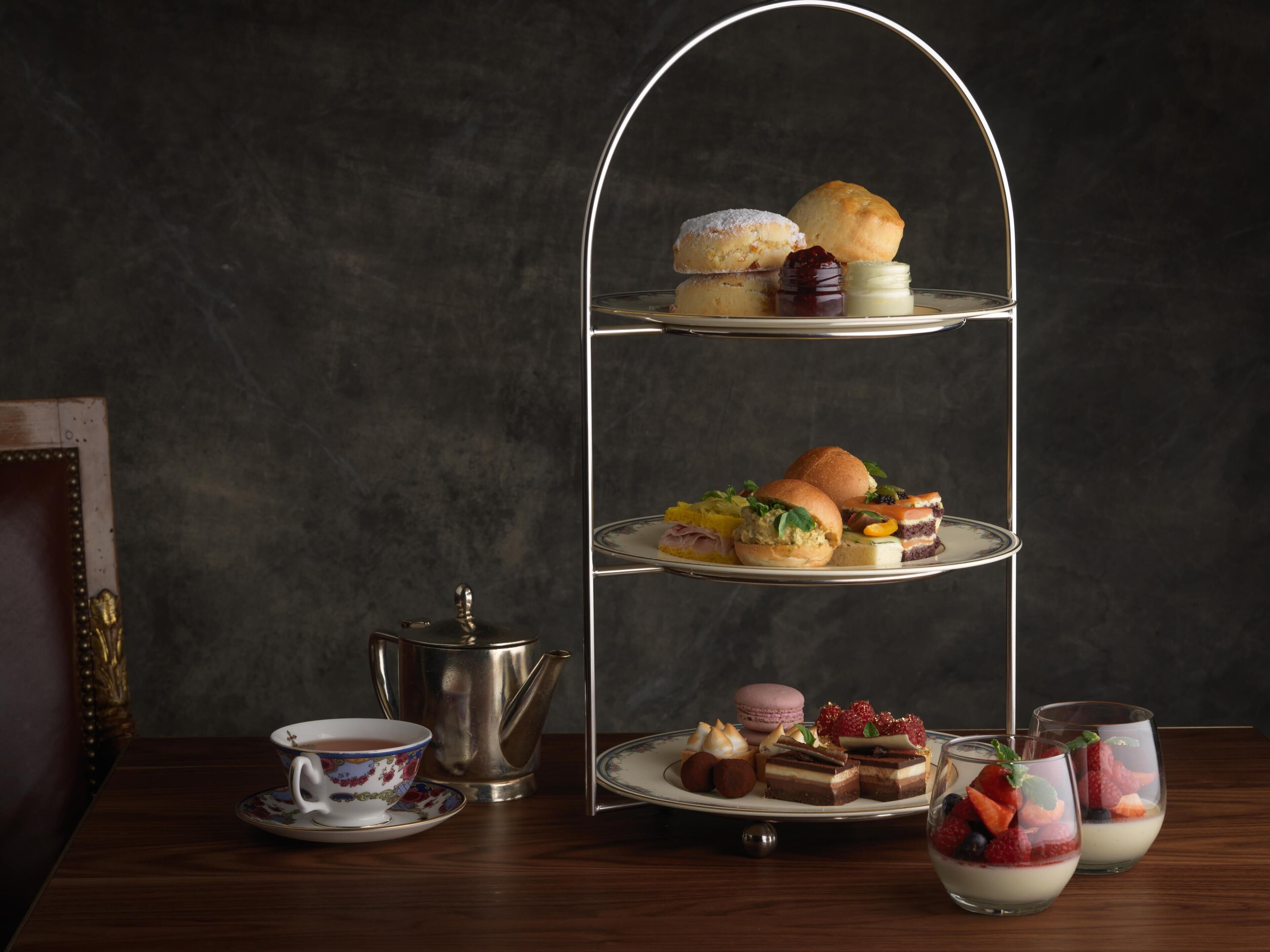 A three-tiered dessert stand with various pastries and desserts, a teapot, and a cup of tea on a wooden table against a dark background.