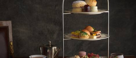 Un soporte de tres niveles con diversos pasteles y postres, una tetera y una taza de té sobre una mesa de madera contra un fondo oscuro.