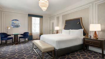 Habitación de hotel con una cama grande, dos mesitas de noche, un banco, una mesa redonda con sillas y una araña.