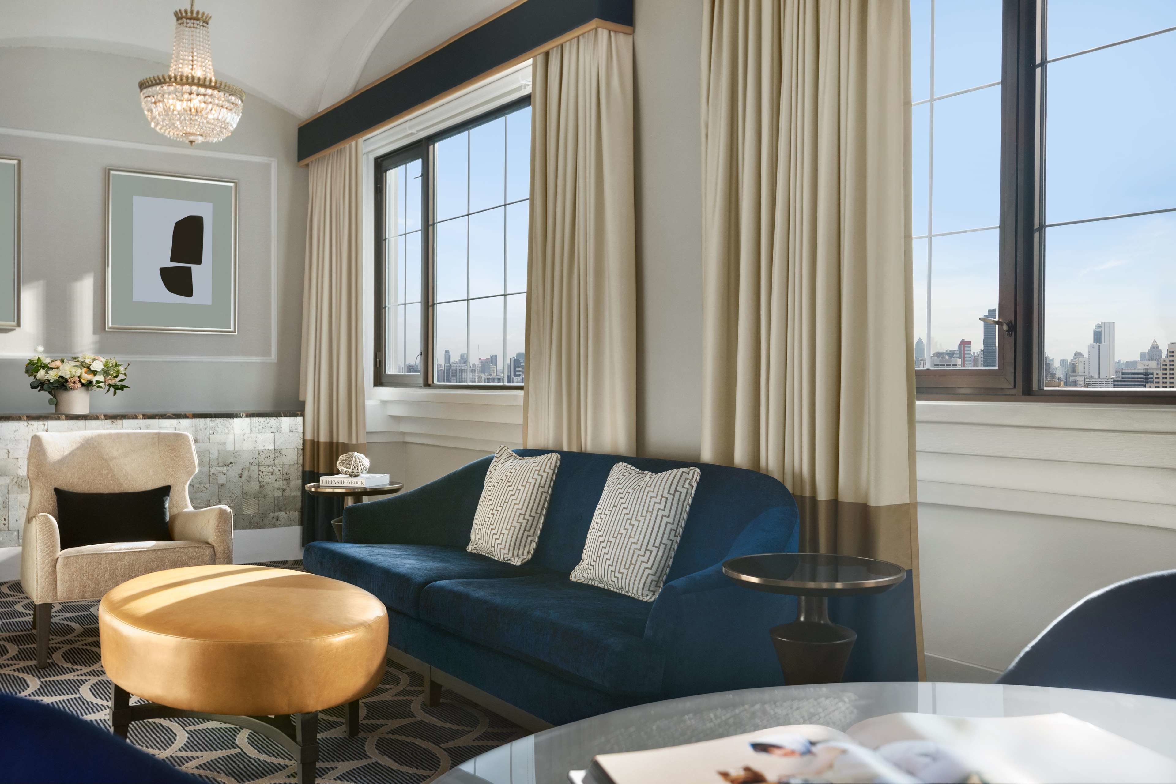 A modern hotel room with a blue sofa, a round ottoman, a small side table, and a large window offering a city view.