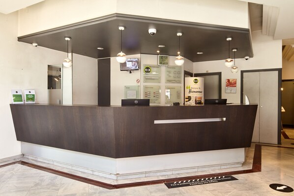 A reception desk with a dark wood finish, multiple light fixtures, a digital screen, and informational signs. - B&B HOTEL Saint-Quentin-en-Yvelines Centre Gare (Montigny-le-Bretonneux)