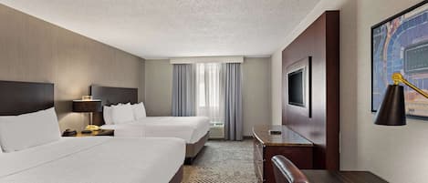 A hotel room with two beds, a desk, a chair, a TV, and a large window with curtains.