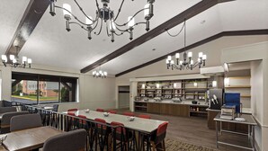 A spacious dining area with a long table, multiple chairs, and a chandelier. There is a kitchen area with a counter and various appliances.