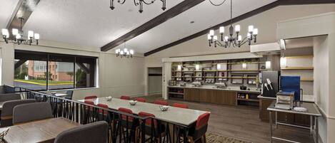 A spacious dining area with a long table, multiple chairs, and a chandelier. There is a kitchen area with a counter and various appliances.