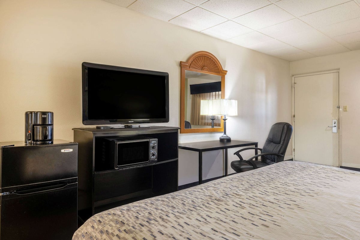 Habitación de hotel con cama, televisión, cafetera, microondas, escritorio, silla y espejo.
