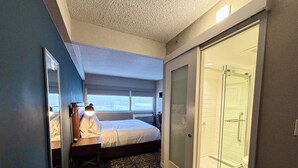 A hotel room with a bed, a desk with a lamp, a walk-in closet, and a glass shower. - Four Points By Sheraton Norwood (Norwood)