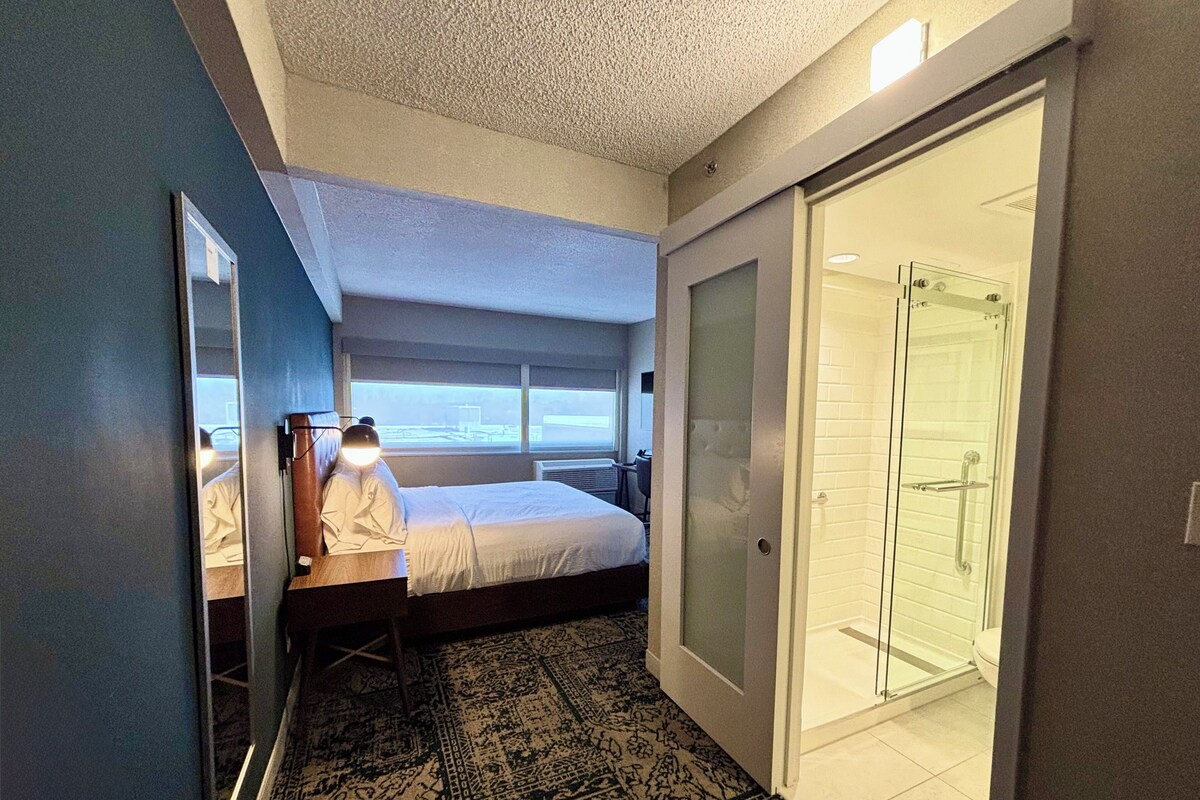 A hotel room with a bed, a desk with a lamp, a walk-in closet, and a glass shower.