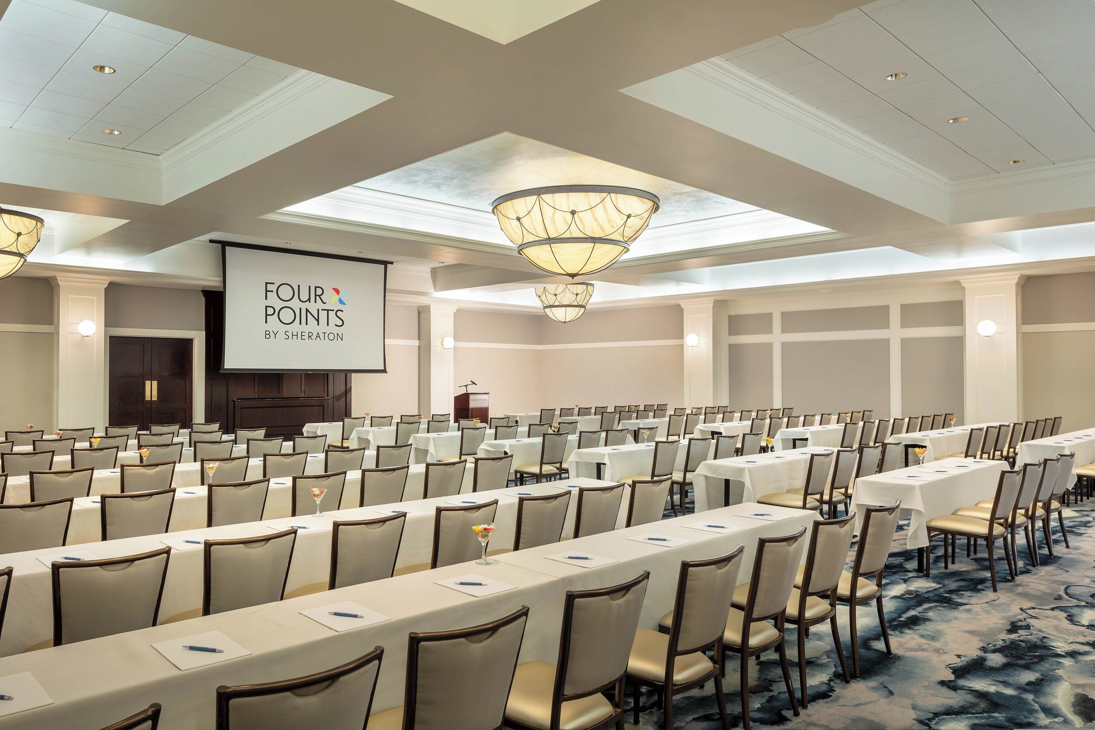 A conference room with rows of tables and chairs, a large screen displaying "FOUR POINTS BY SHERATON", and a ceiling with skylights and chandeliers.