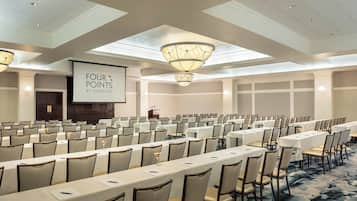 A conference room with rows of tables and chairs, a large screen displaying "FOUR POINTS BY SHERATON", and a ceiling with skylights and chandeliers.