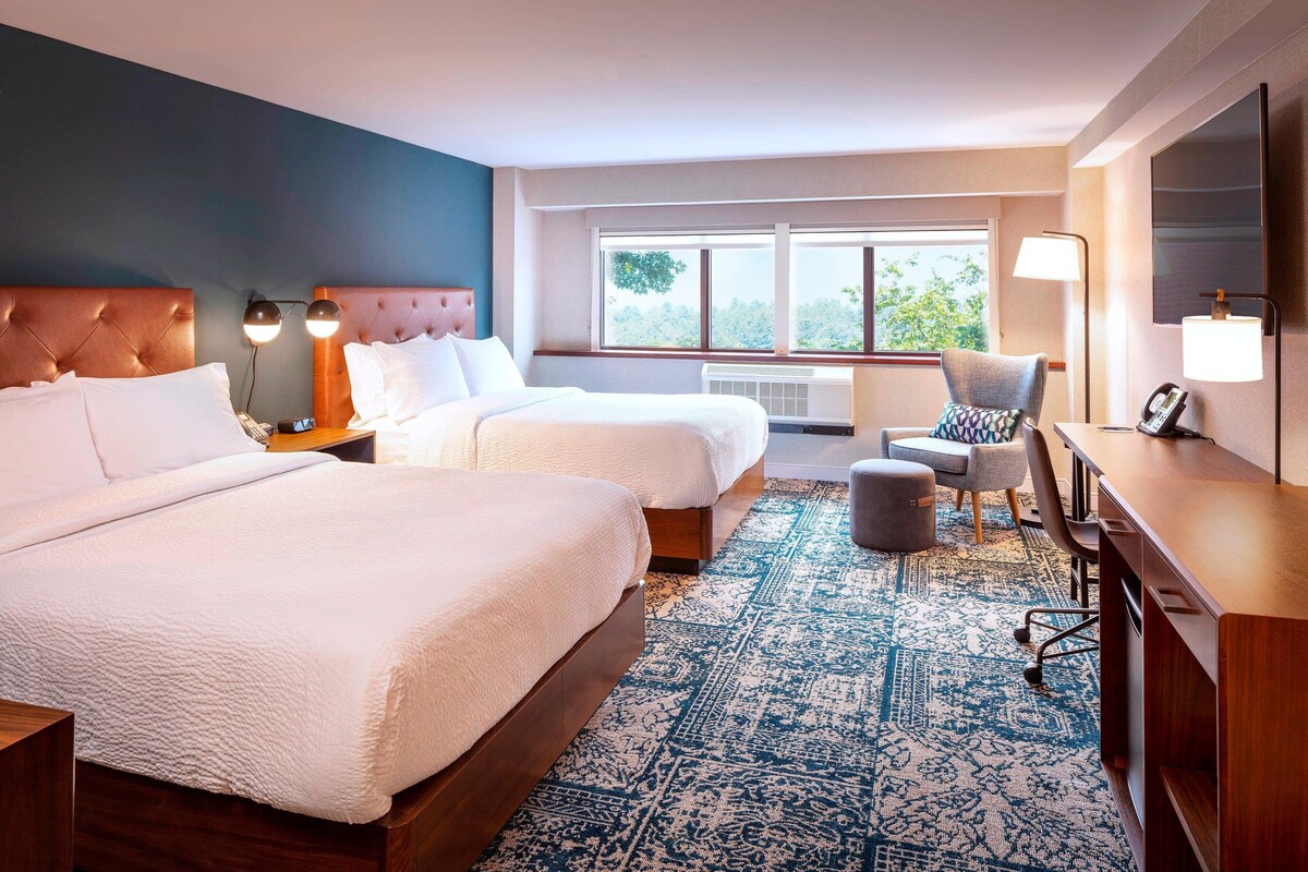 A hotel room with two beds, a desk, a chair, a TV, and a large window with a view of trees.