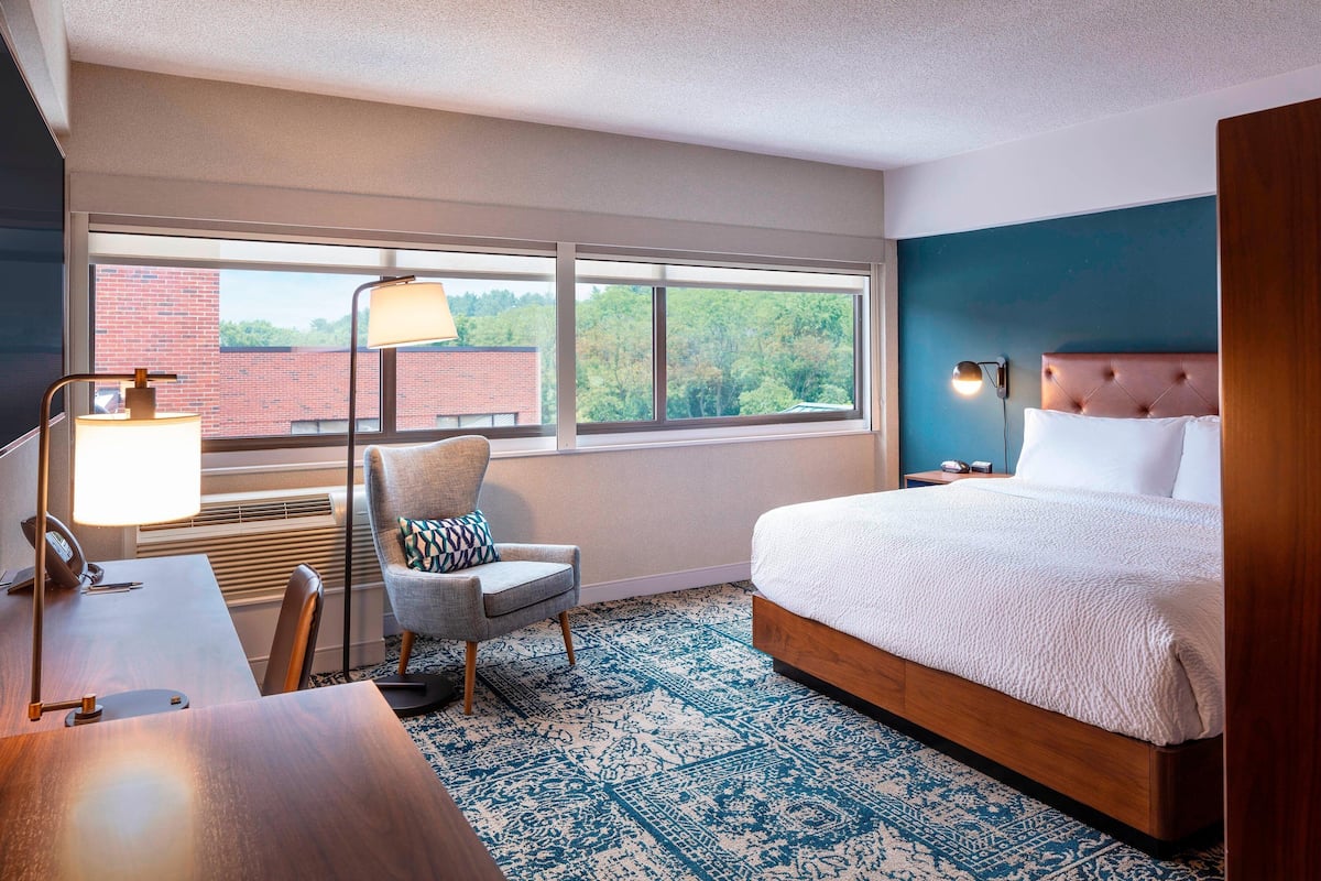 A hotel room with a large bed, a desk with a lamp, a chair, and a view of a brick building through a large window.