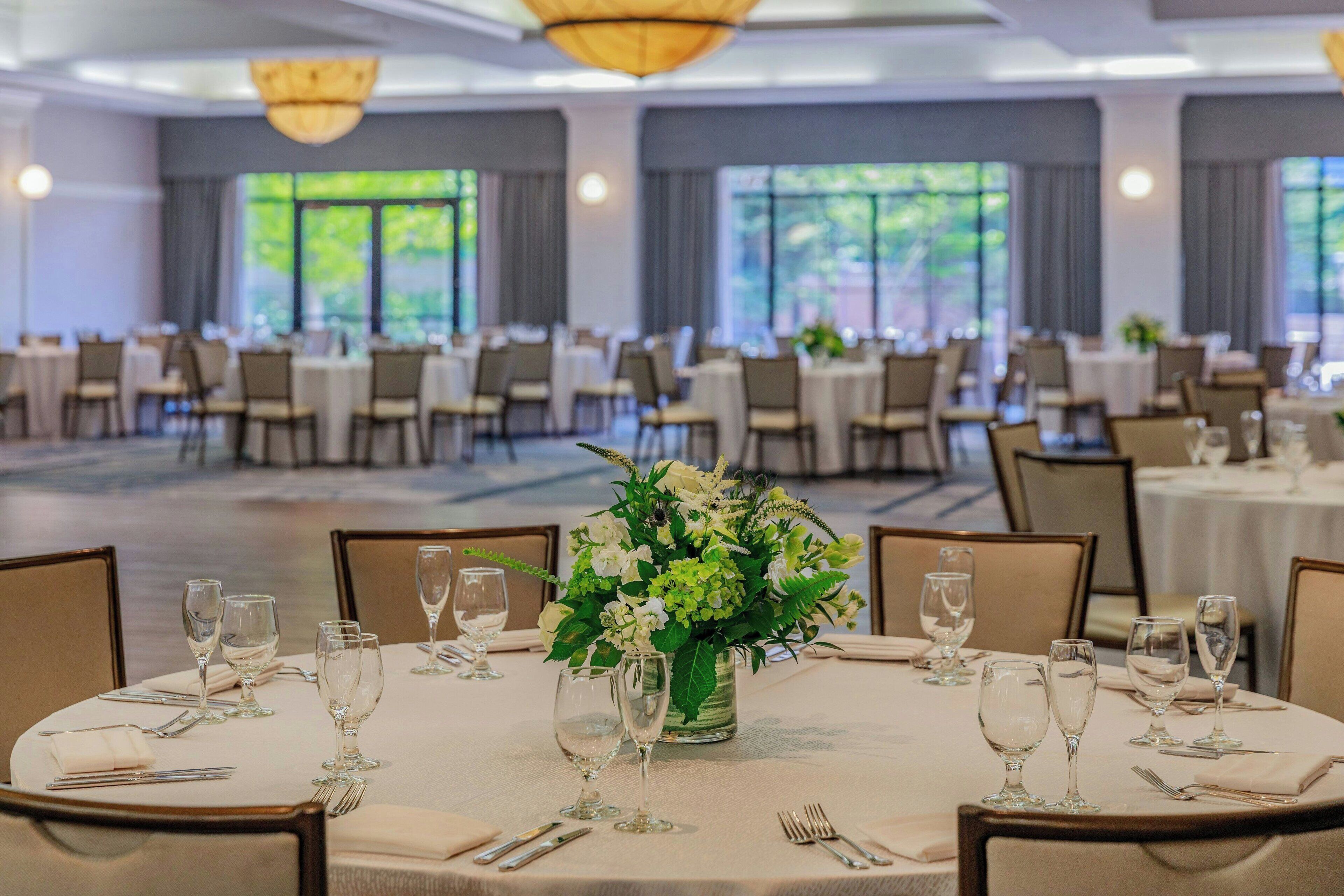 Une salle de banquet avec des tables rondes dressées pour le repas, chacune agrémentée d’un centre de table composé de fleurs et de verres transparents.