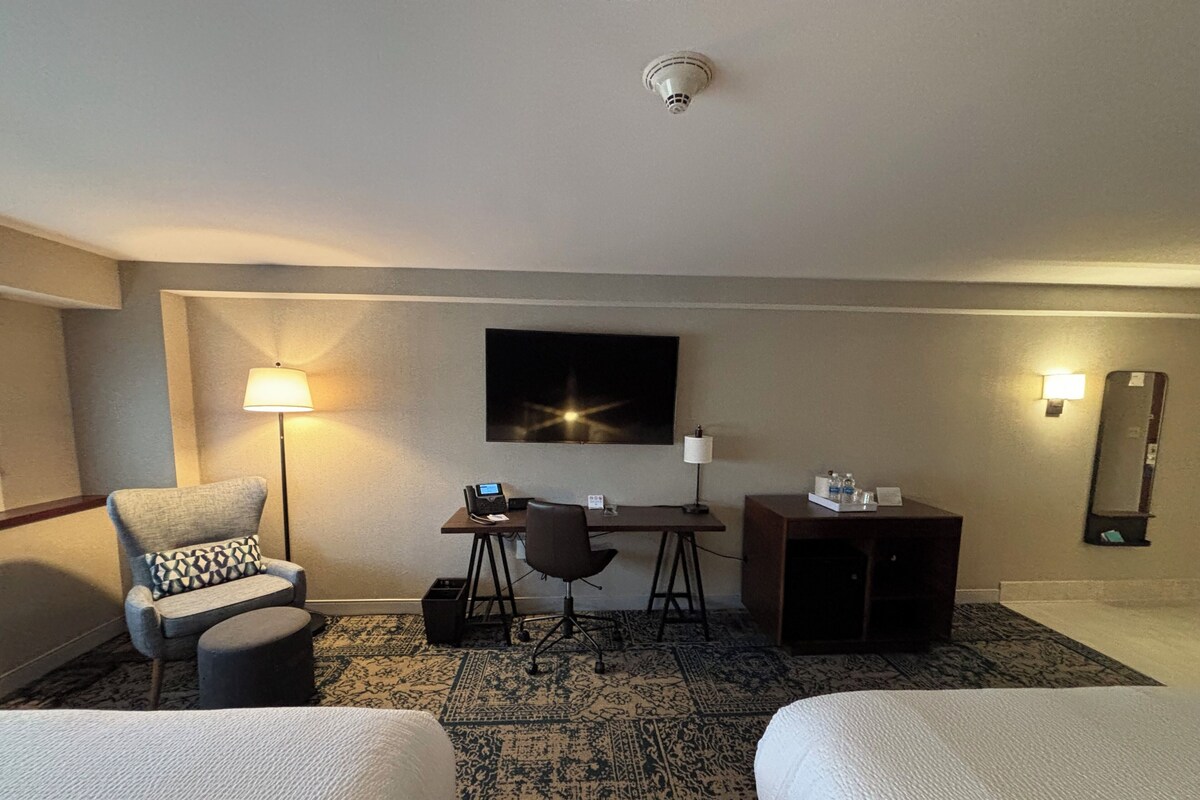 A hotel room with two beds, a desk with a computer, a chair, a lamp, a small cabinet, and a wall-mounted TV.