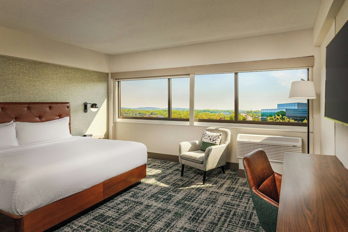 A hotel room with a large bed, a chair, a desk, a window with a view, and a wall-mounted lamp.