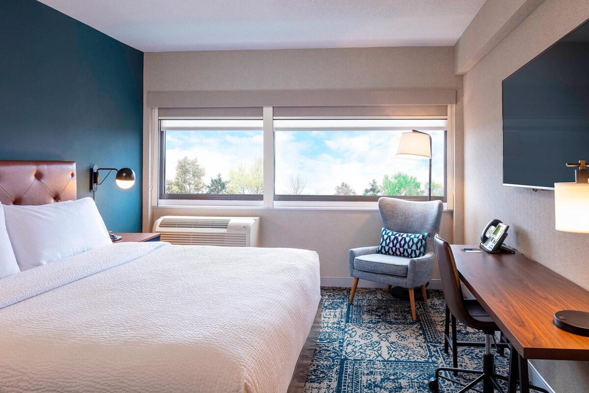A hotel room with a bed, a desk, a chair, a large window, and a wall-mounted TV.