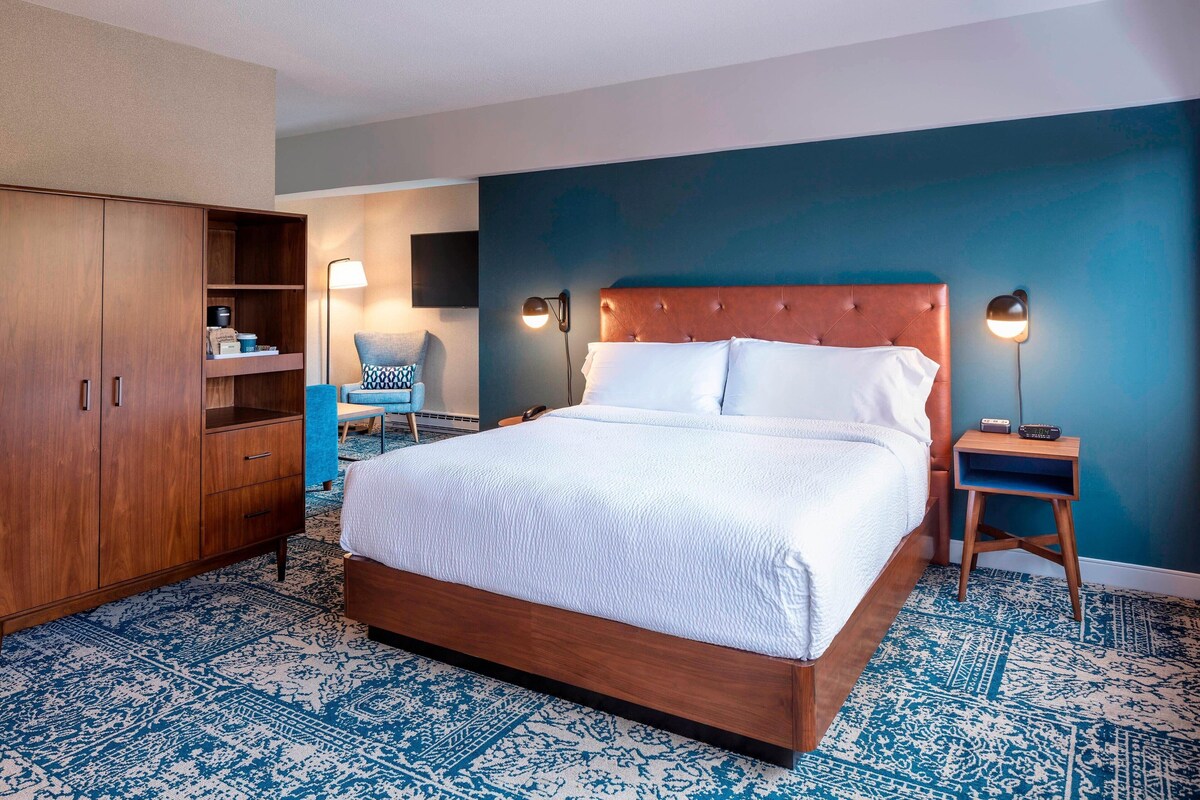 A bedroom with a large bed, a wardrobe, a chair, a desk, and a wall-mounted TV.