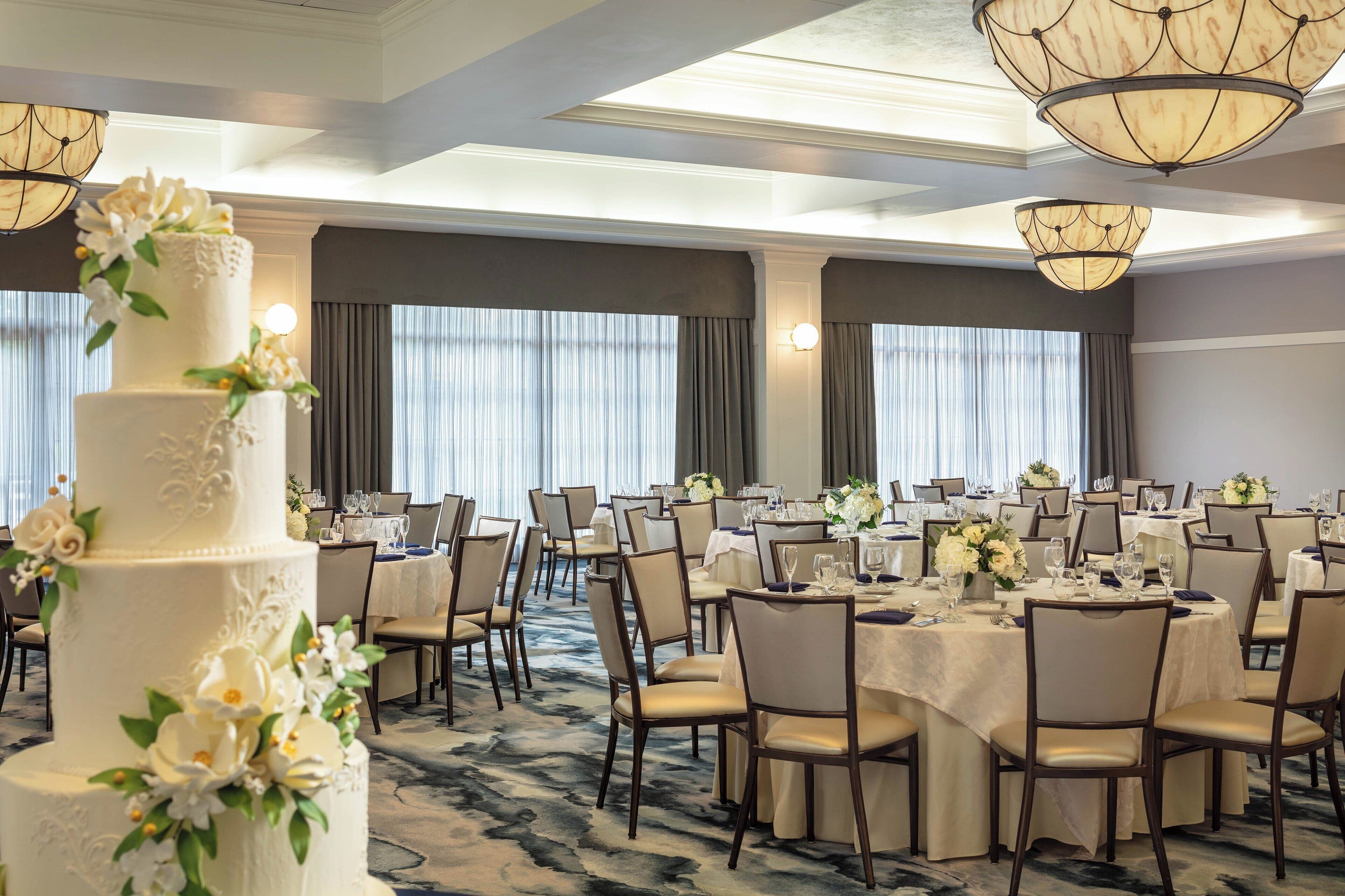 Una sala de banquets amb taules rodones, cadirees i un pastís blanc gran decorat amb flors.