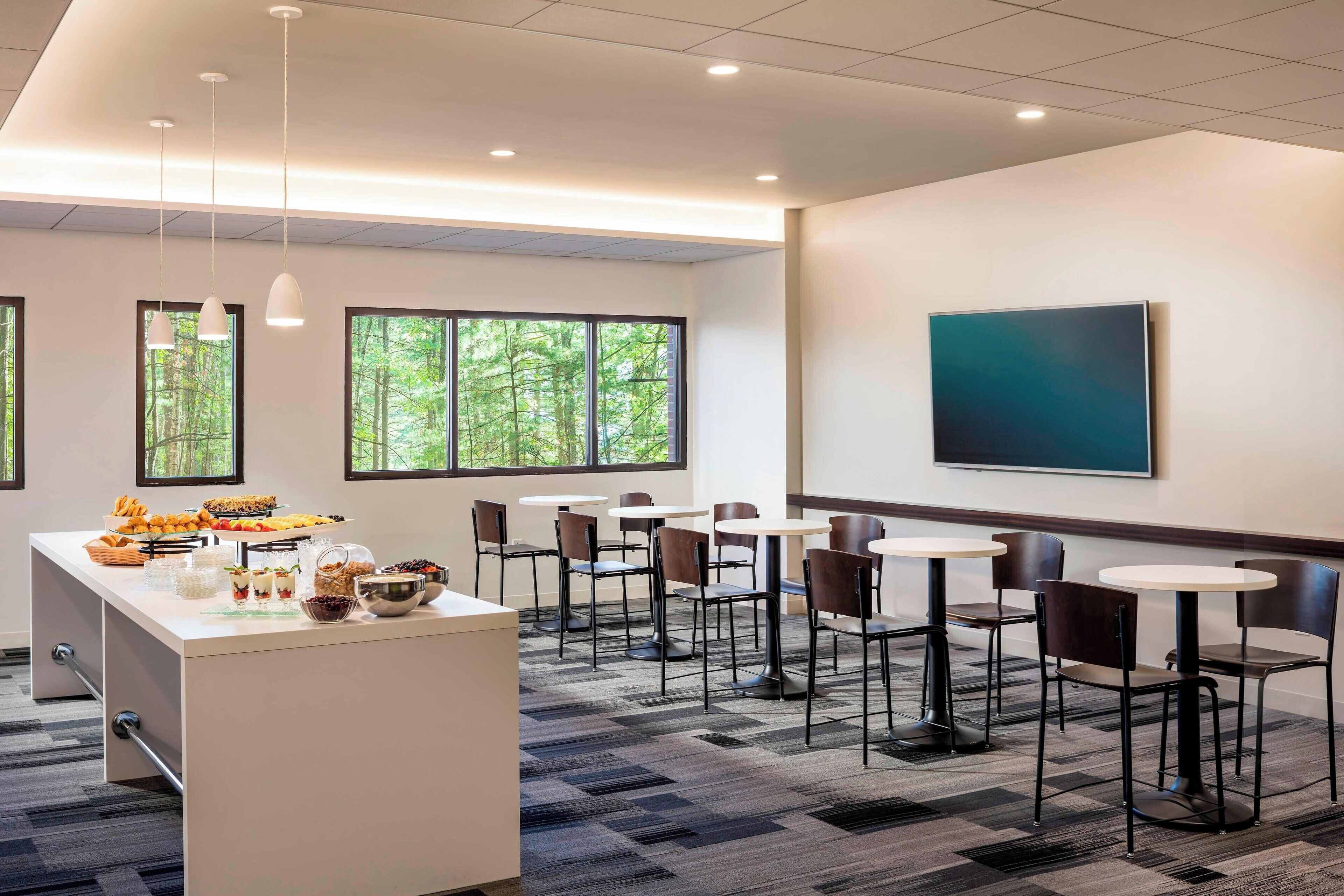 A modern hotel room with a breakfast bar, multiple seating areas, and large windows offering a view of greenery.