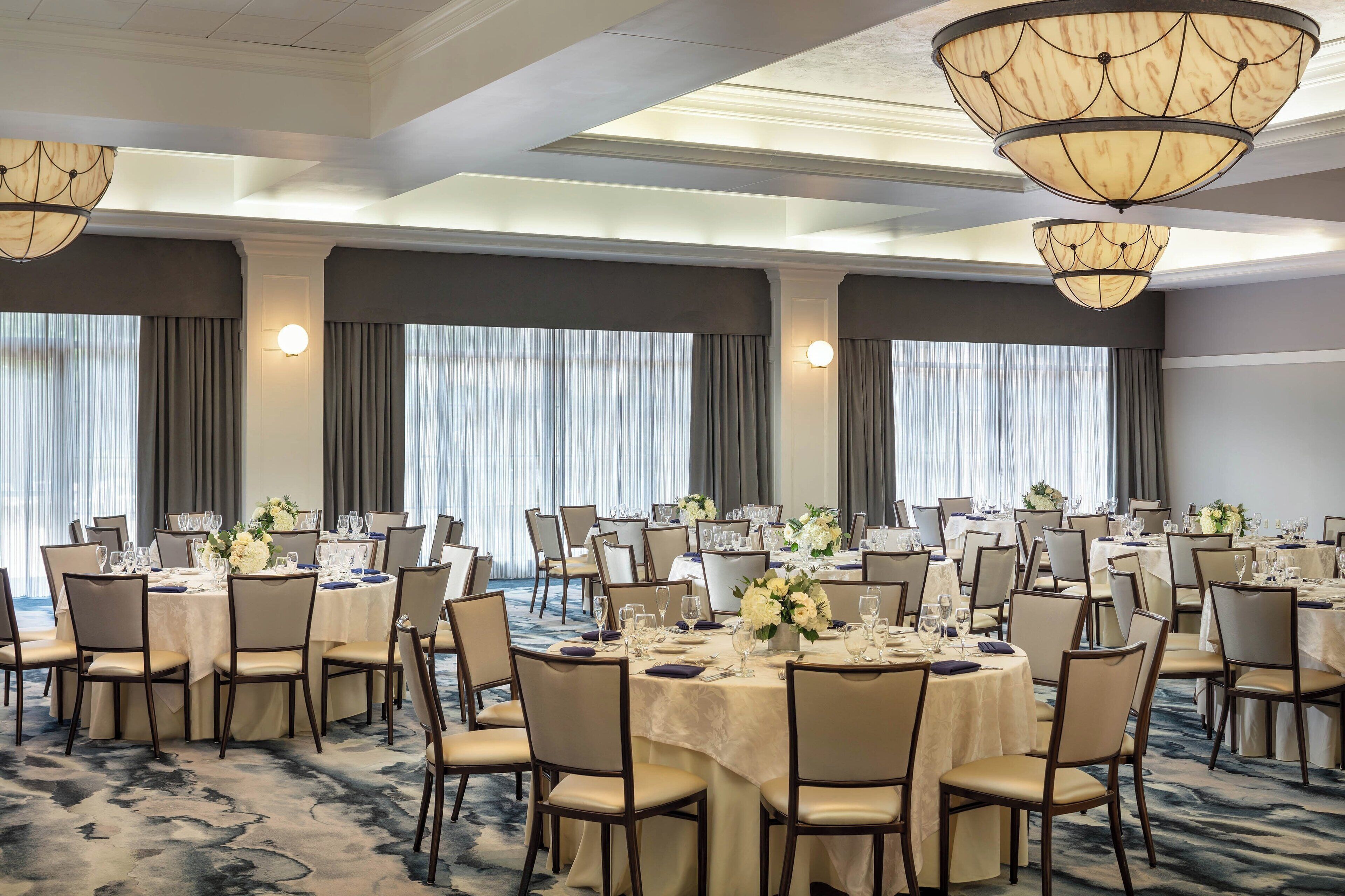 A banquet hall with round tables set for dining, large windows with curtains, and chandeliers.