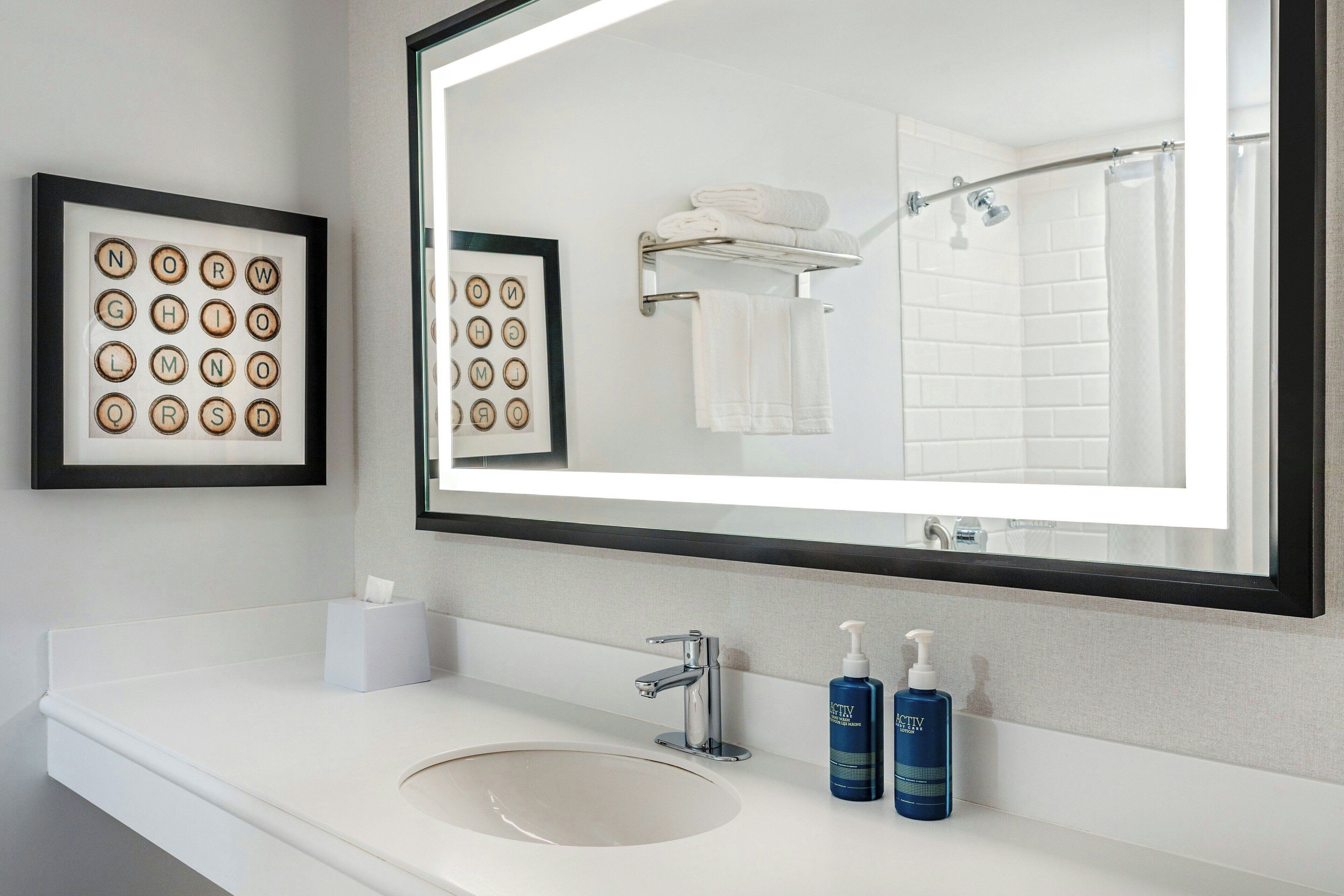 A bathroom with a large mirror, a sink, two bottles of lotion, and framed word puzzles on the wall.