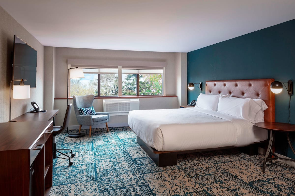 A hotel room with a large bed, a desk with a chair, a TV, and a window with a view of trees.