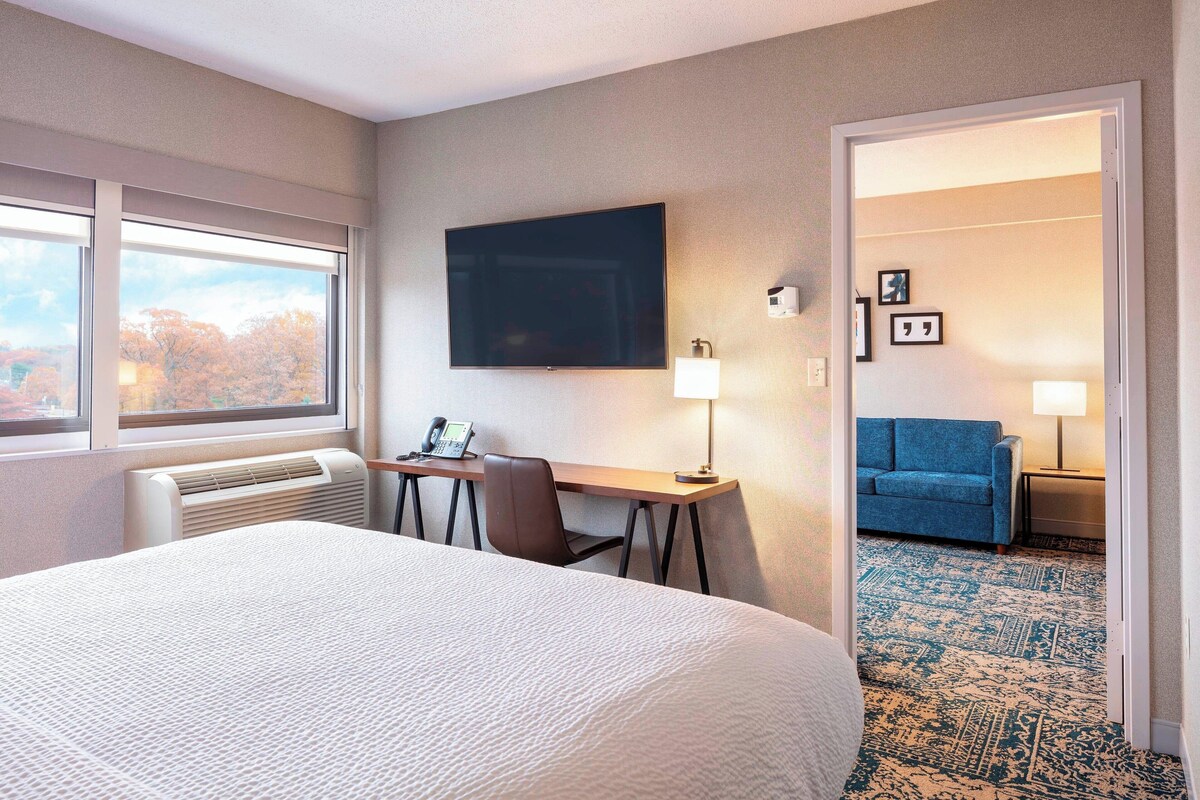 A hotel room with a large bed, a desk with a computer, a TV mounted on the wall, and a view of trees through the window.