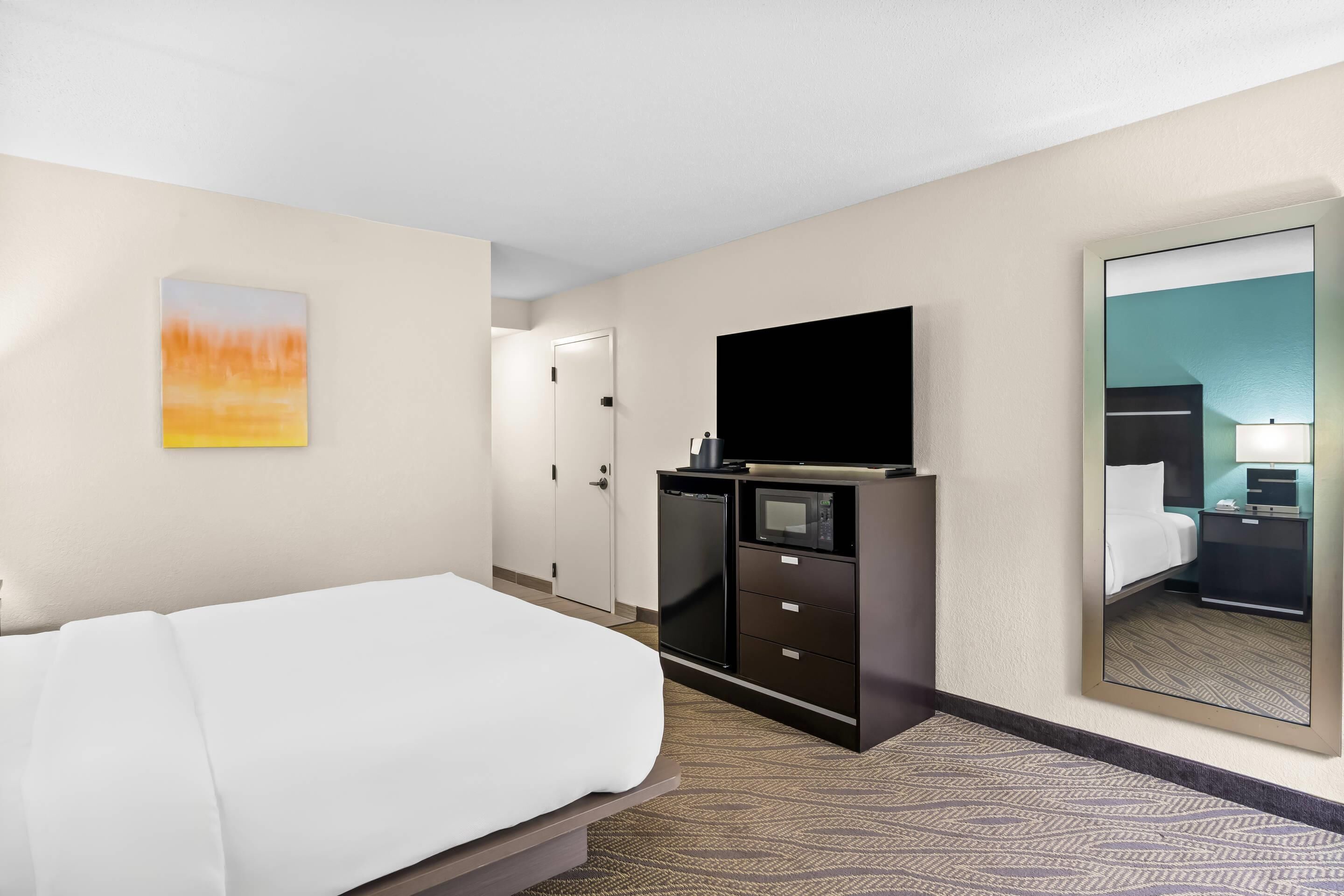 Una habitación de hotel con cama, televisión, minibar y un espejo que refleja el ambiente.