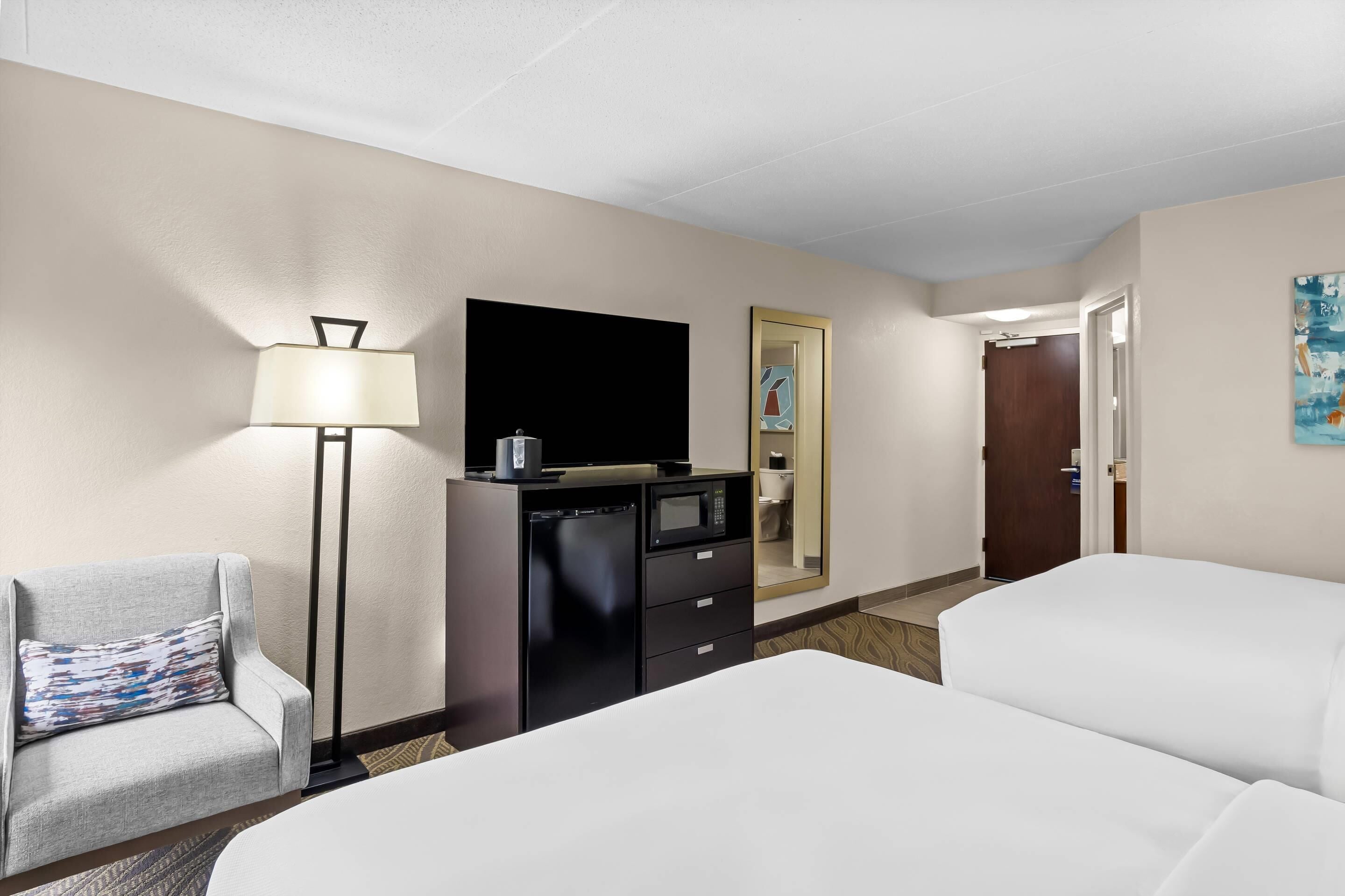 A hotel room with two beds, a desk with a lamp, a TV, a microwave, and a dresser.
