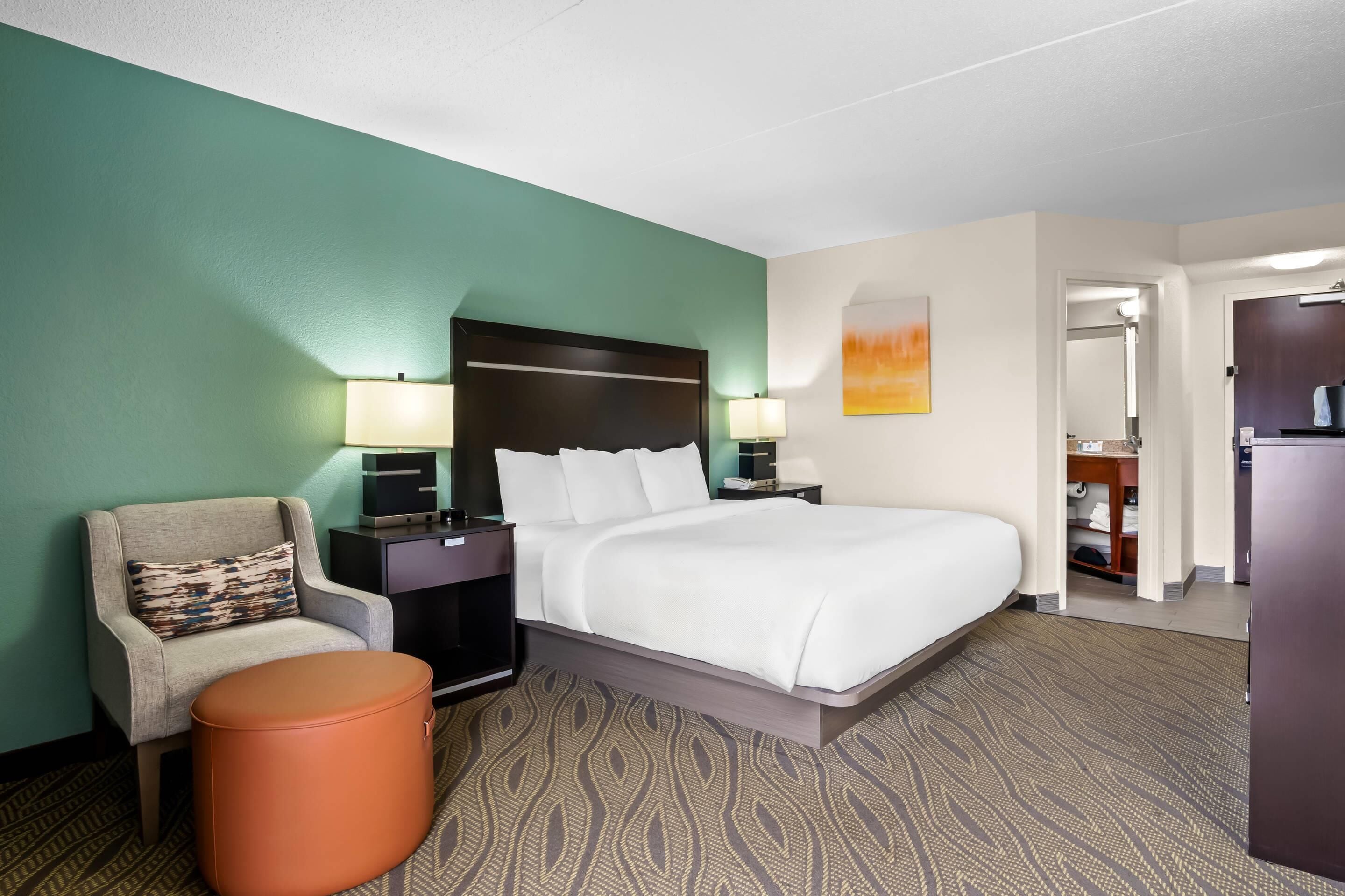 A hotel room with a bed, a chair, a nightstand, a lamp, and a small ottoman.