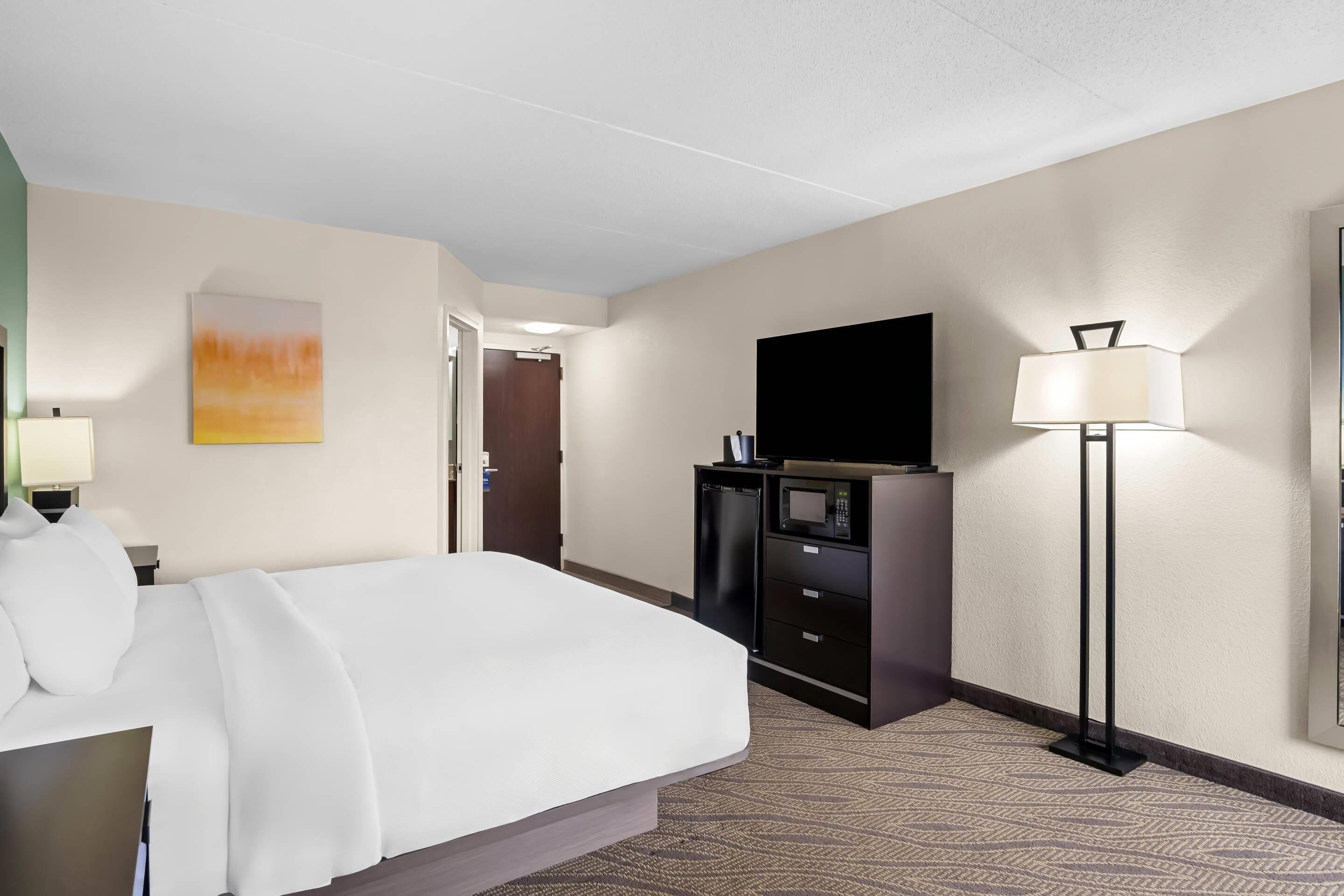 A hotel room with a bed, a flat-screen TV, a desk with a lamp, a microwave, and a small refrigerator.